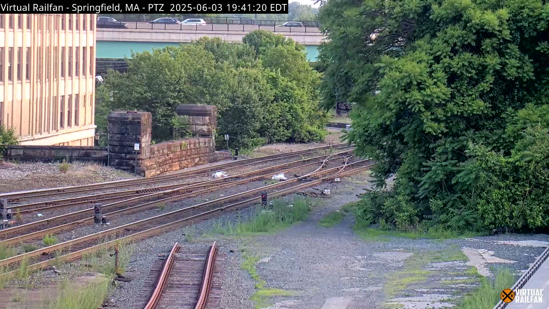 Multiple sets of train tracks intersect near a stone structure and lush green trees under a partly cloudy sky.