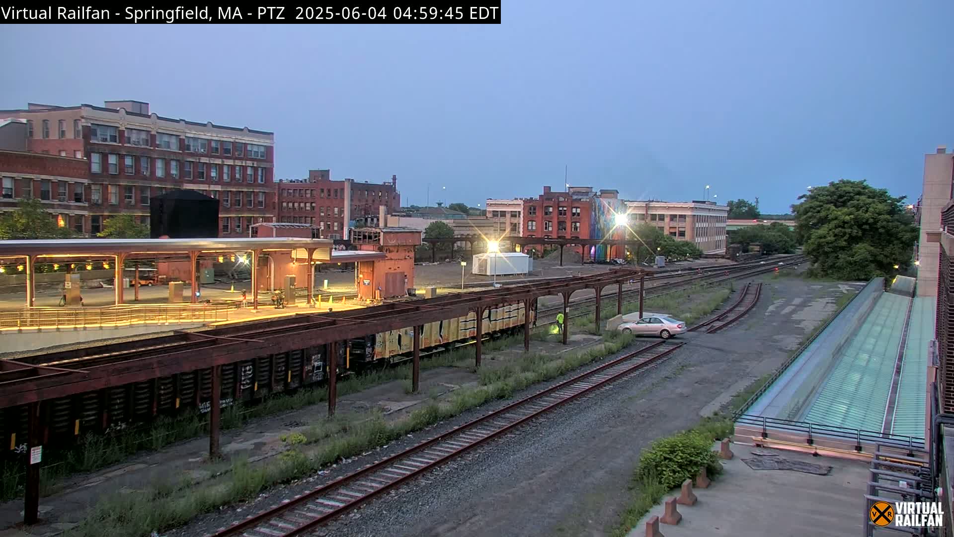 A train station with several railway tracks, a few buildings, and a single car is visible under a twilight sky.