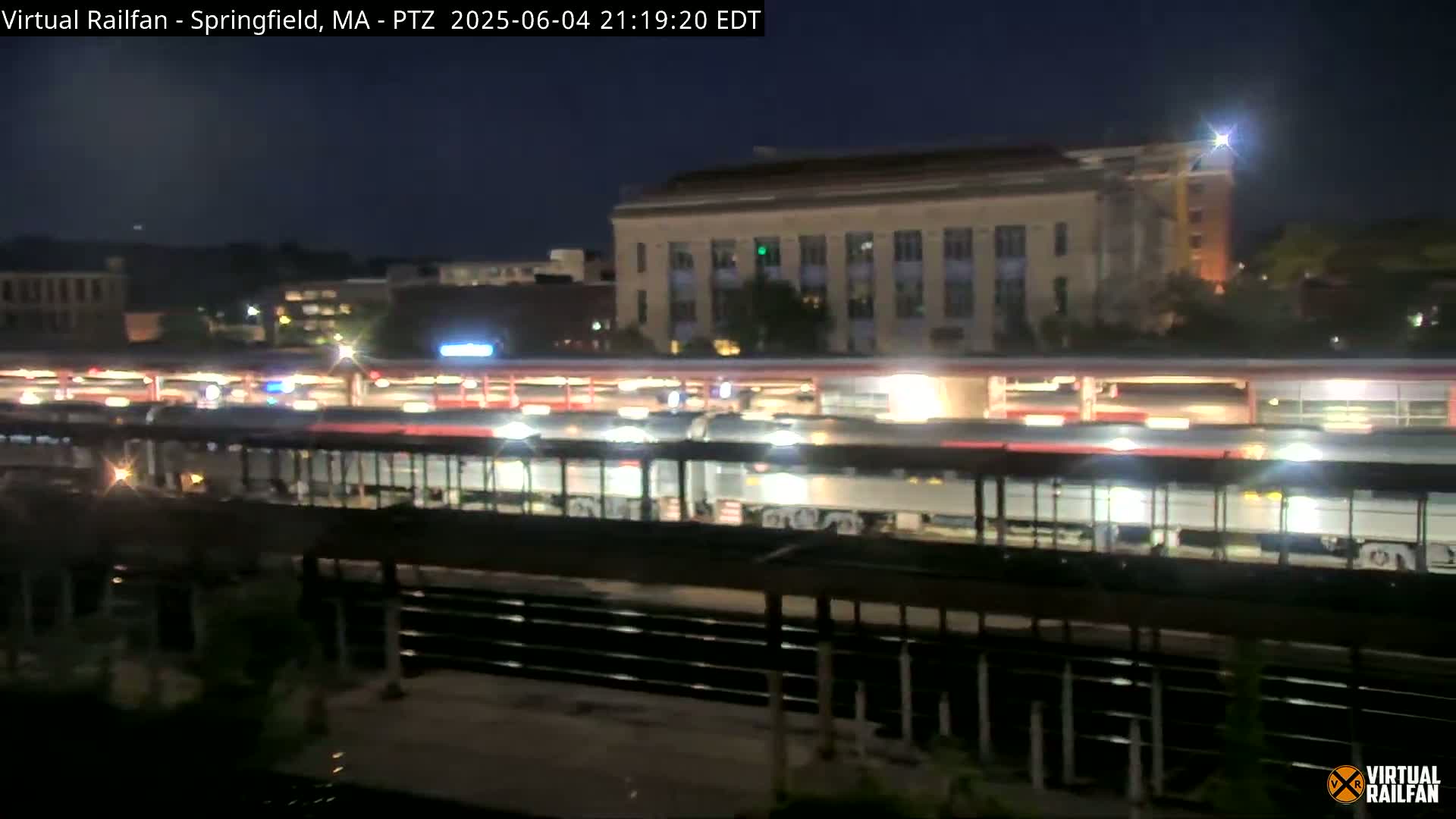 A nighttime view of a train at a station, with a large building visible in the background under a clear sky.
