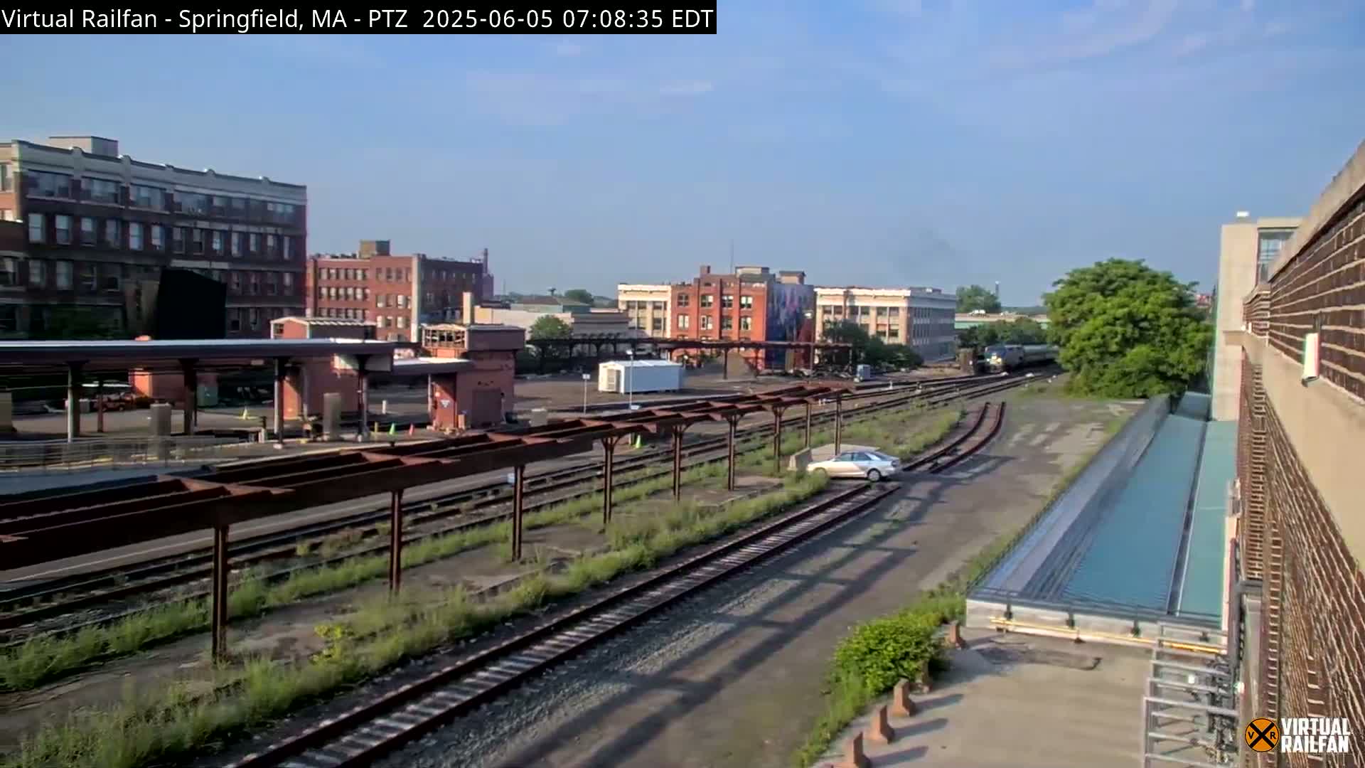 A high-angle view shows a train station with multiple train tracks, several buildings, a single car, and a mostly clear sky on a sunny day.
