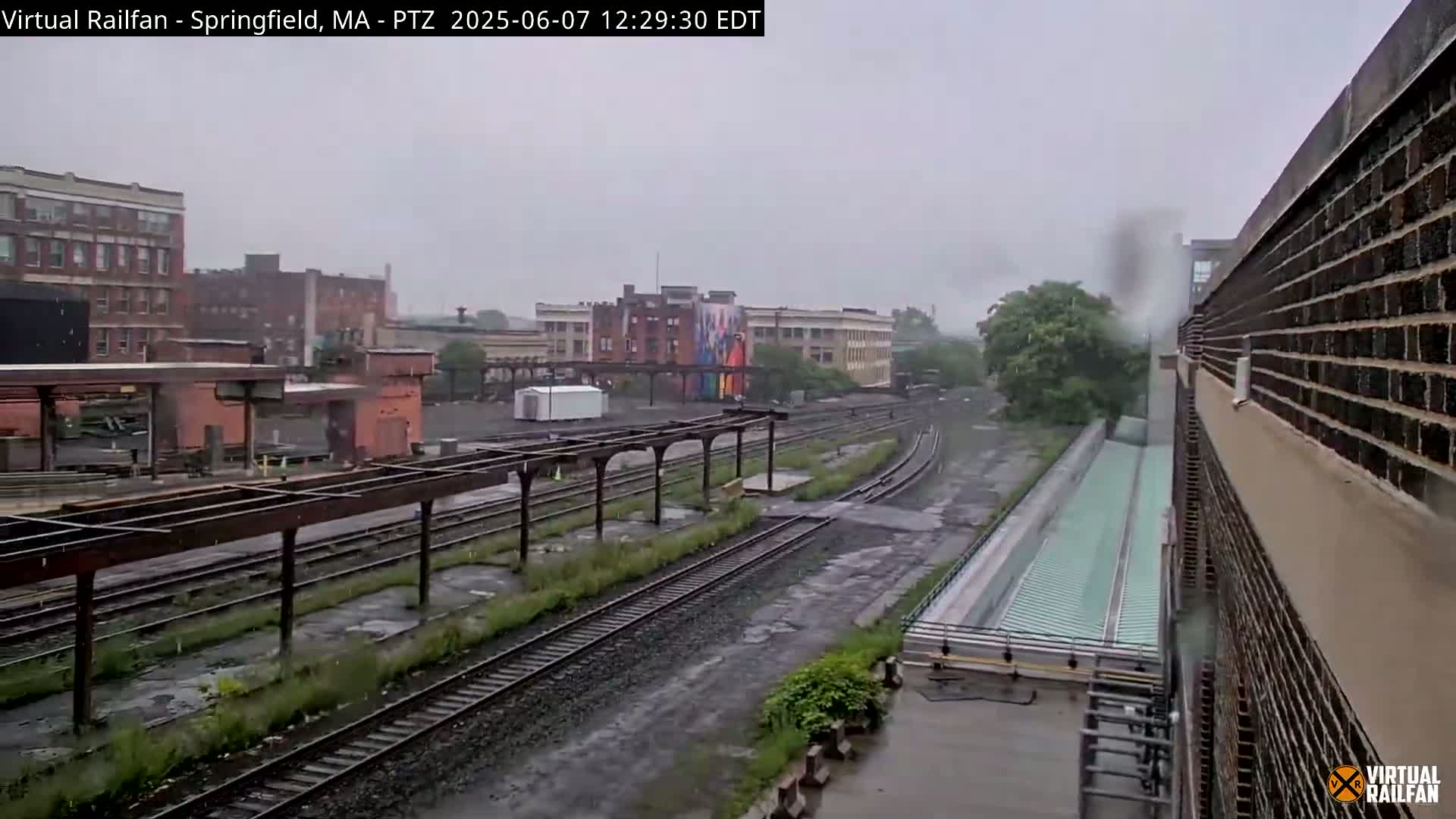 A rainy view of several train tracks and adjacent buildings, including a brick building with a long covered platform.