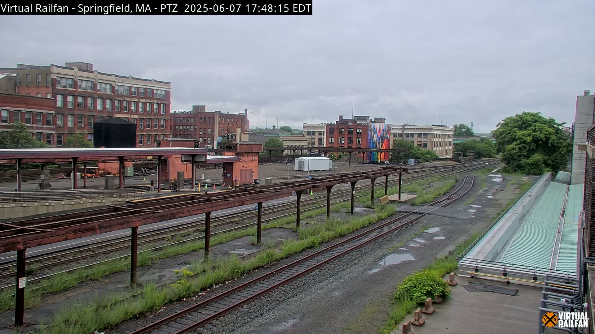 A view of a train yard with several railway tracks, overgrown weeds, and brick buildings under a cloudy sky.
