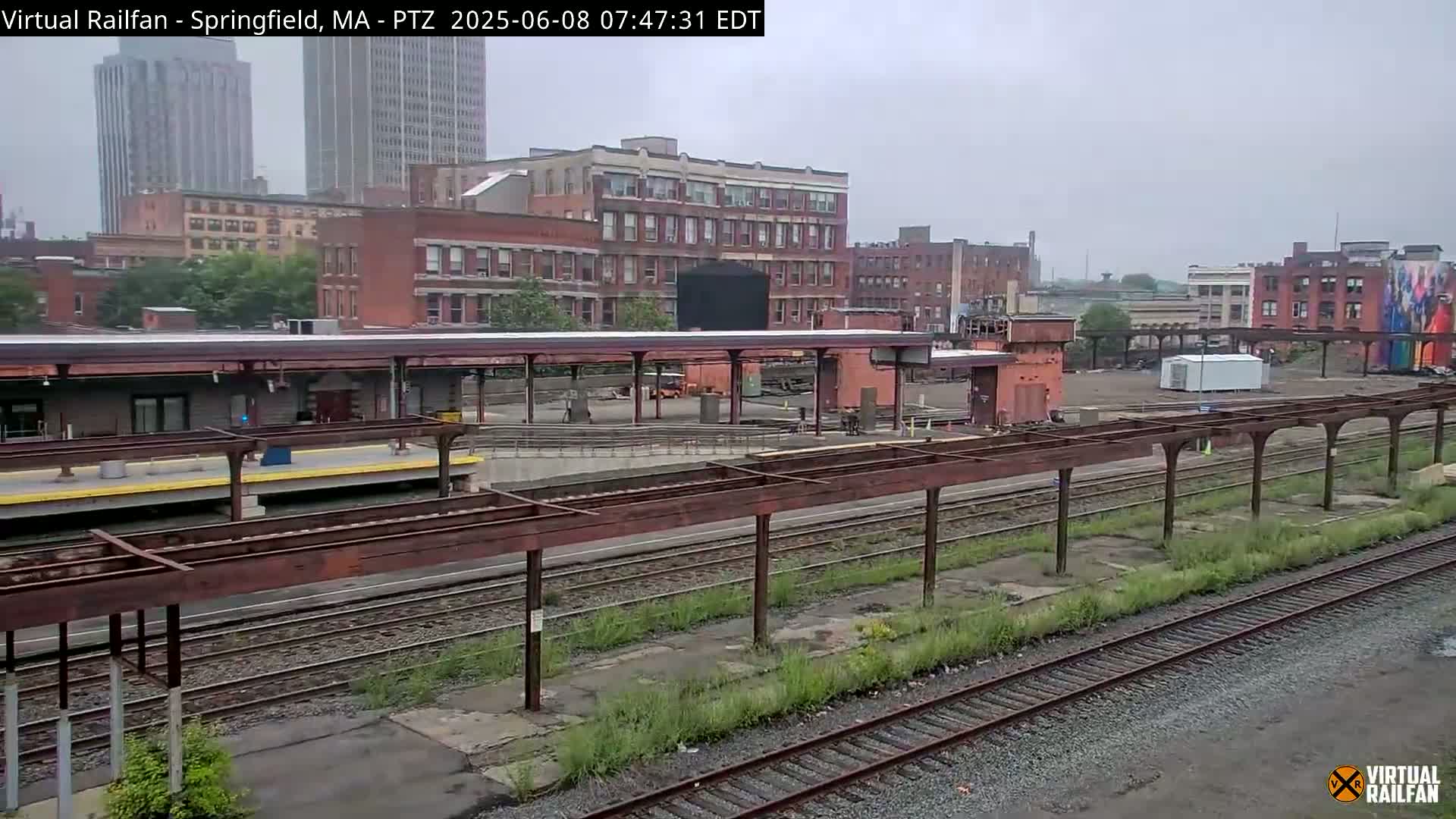 A train station platform with several tracks and overhead shelters, situated amidst brick buildings under an overcast sky.