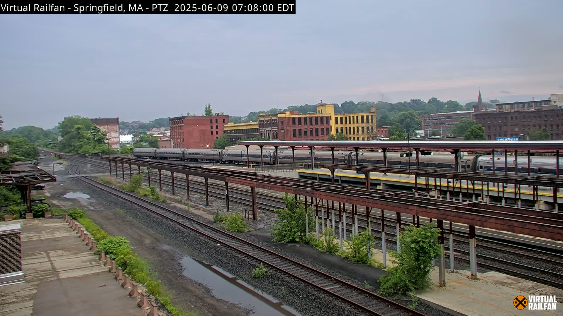 Several passenger trains are at a station under an overcast sky, with industrial buildings and trees in the background.