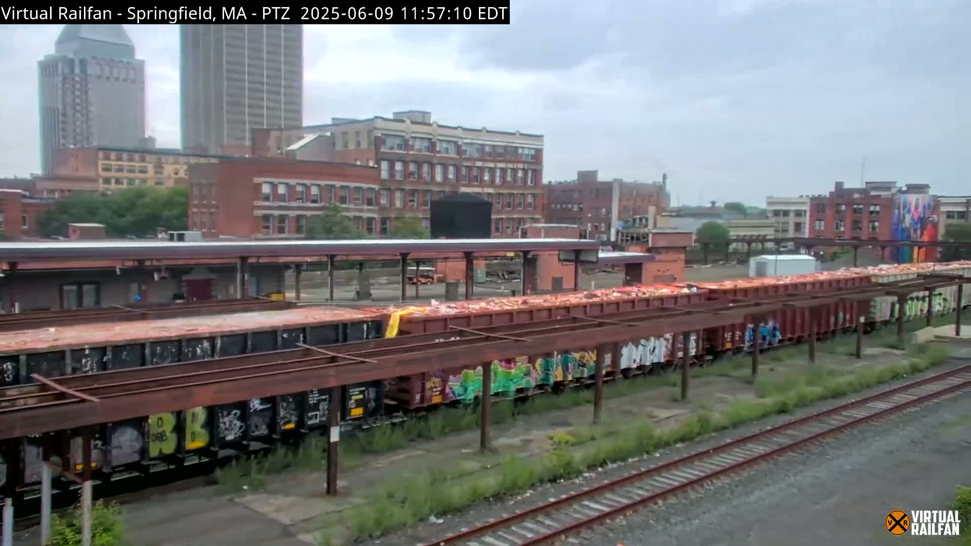 A long freight train filled with what appears to be scrap metal sits under an overcast sky next to a train station in an urban area.