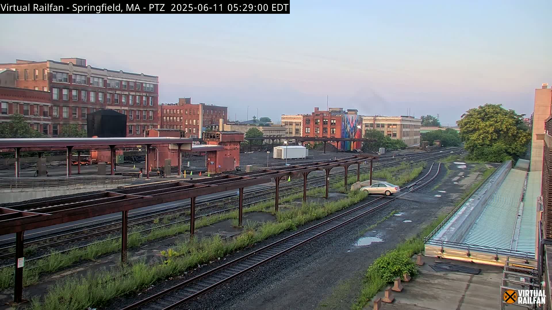 A train yard with multiple train tracks, a platform, and several brick buildings under a clear, light-colored sky.