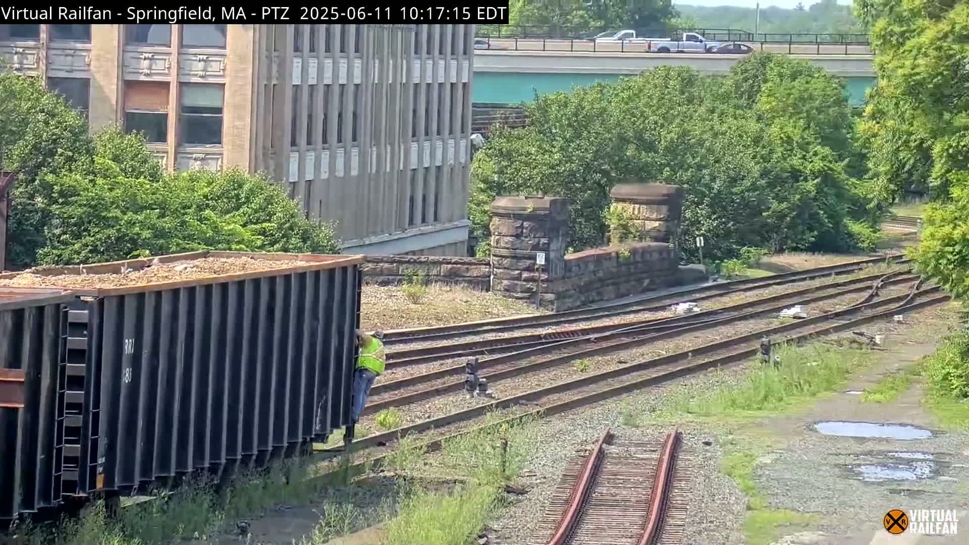 A train car filled with a light brown material sits on railroad tracks next to a stone bridge, under a sunny sky.