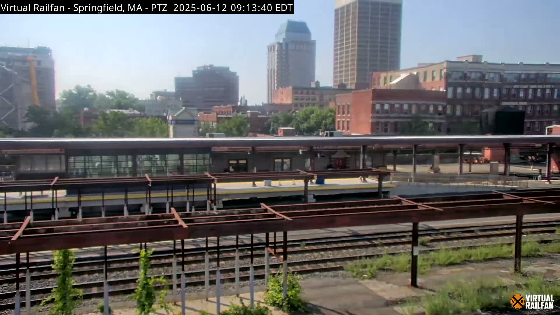A train station platform with several tracks, some buildings and tall skyscrapers in the background on a sunny day.