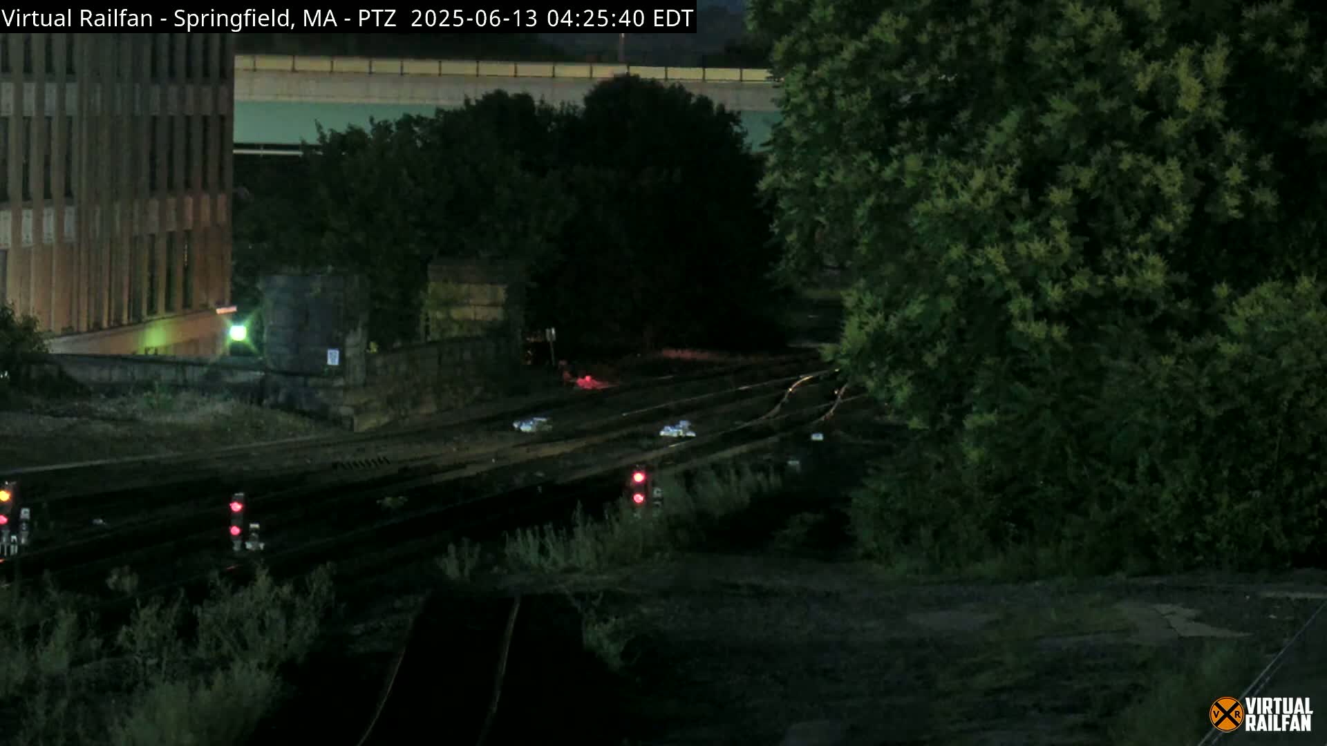 A nighttime view of railroad tracks with several red signal lights, adjacent to a building and some trees under a dark sky.