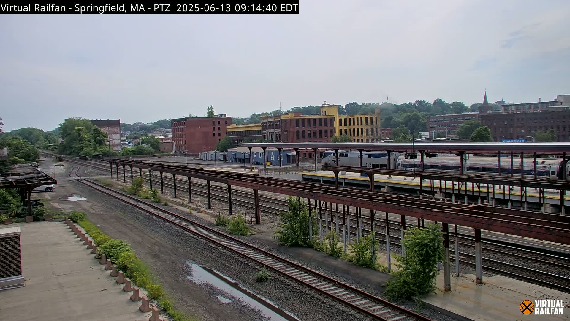 A train sits at a station platform next to several railroad tracks, with industrial buildings and trees visible in the background under an overcast sky.