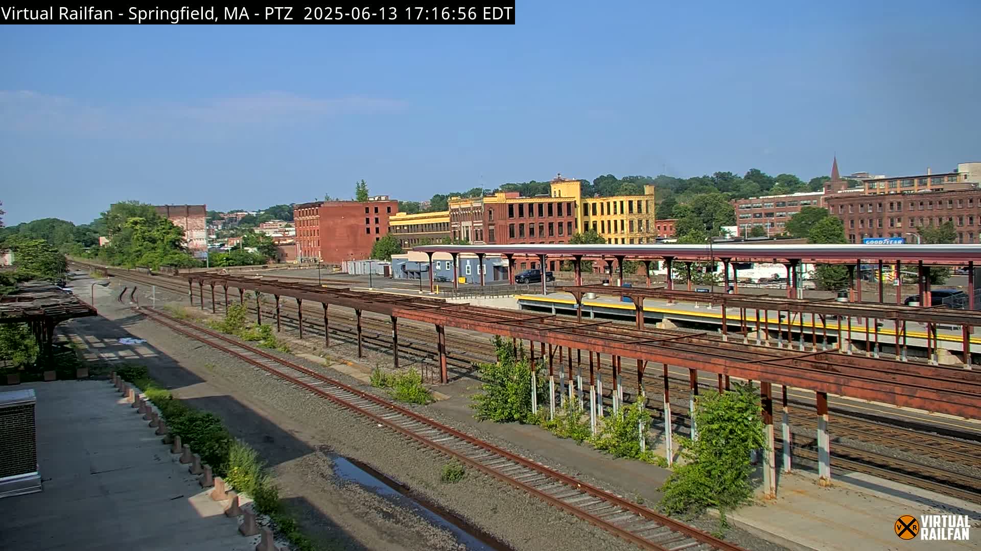 A train station with multiple tracks and rusty overhead shelters is shown under a clear, sunny sky, with several brick buildings in the background.