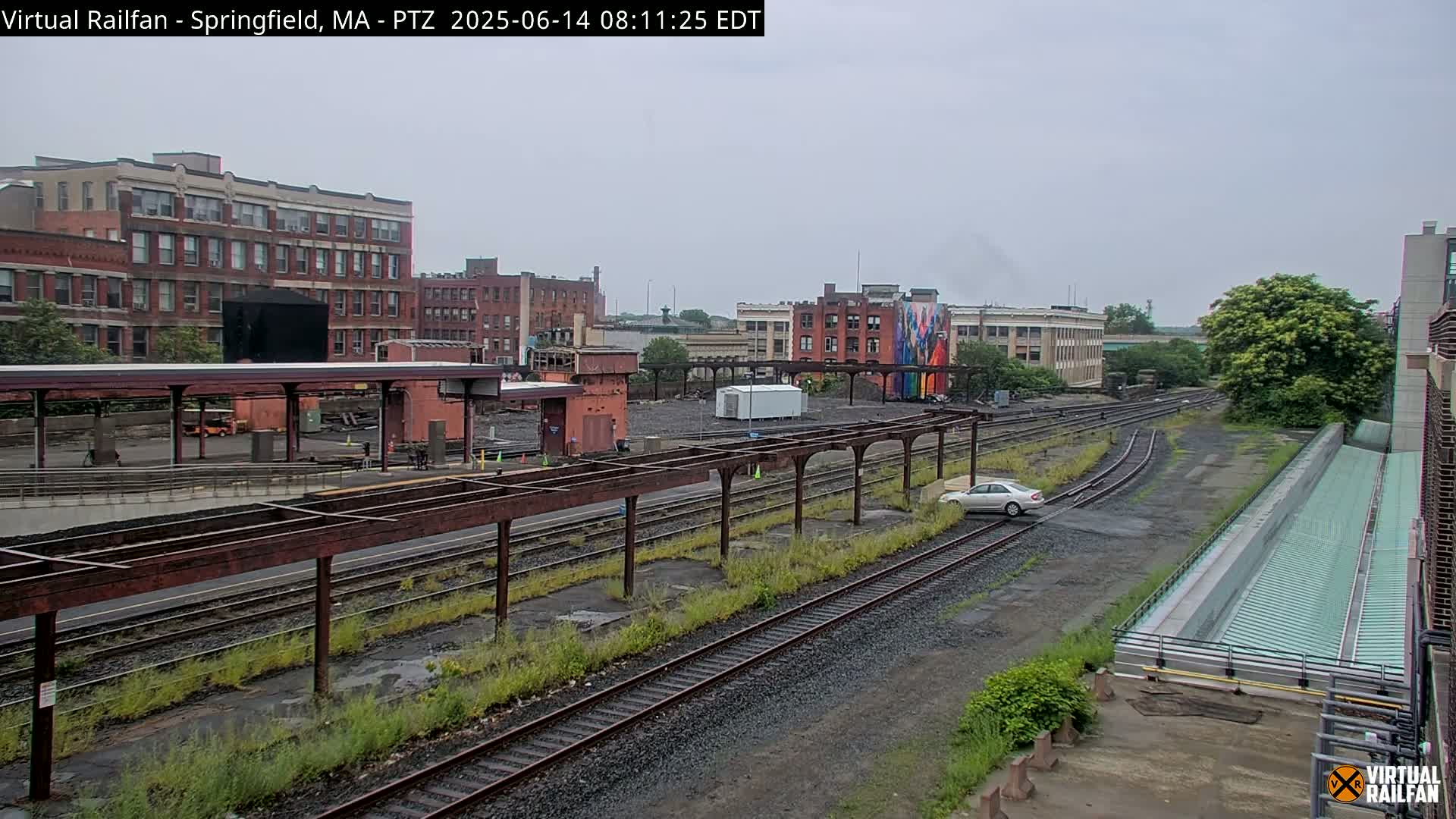 A mostly overcast day reveals a train yard with several railway tracks, industrial buildings, and a single car parked on the grounds.