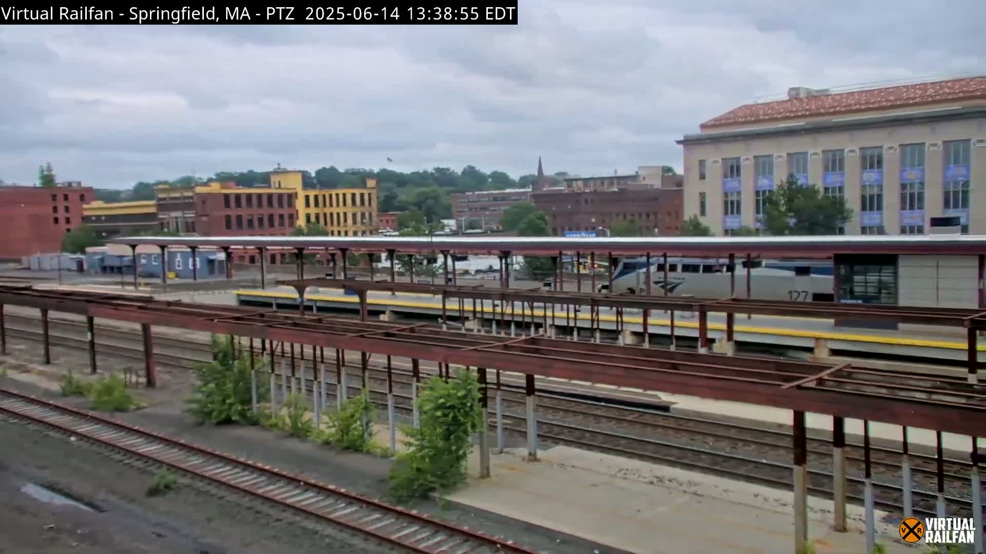 A long, silver train sits at a station under an overcast sky, next to several industrial buildings and train tracks.