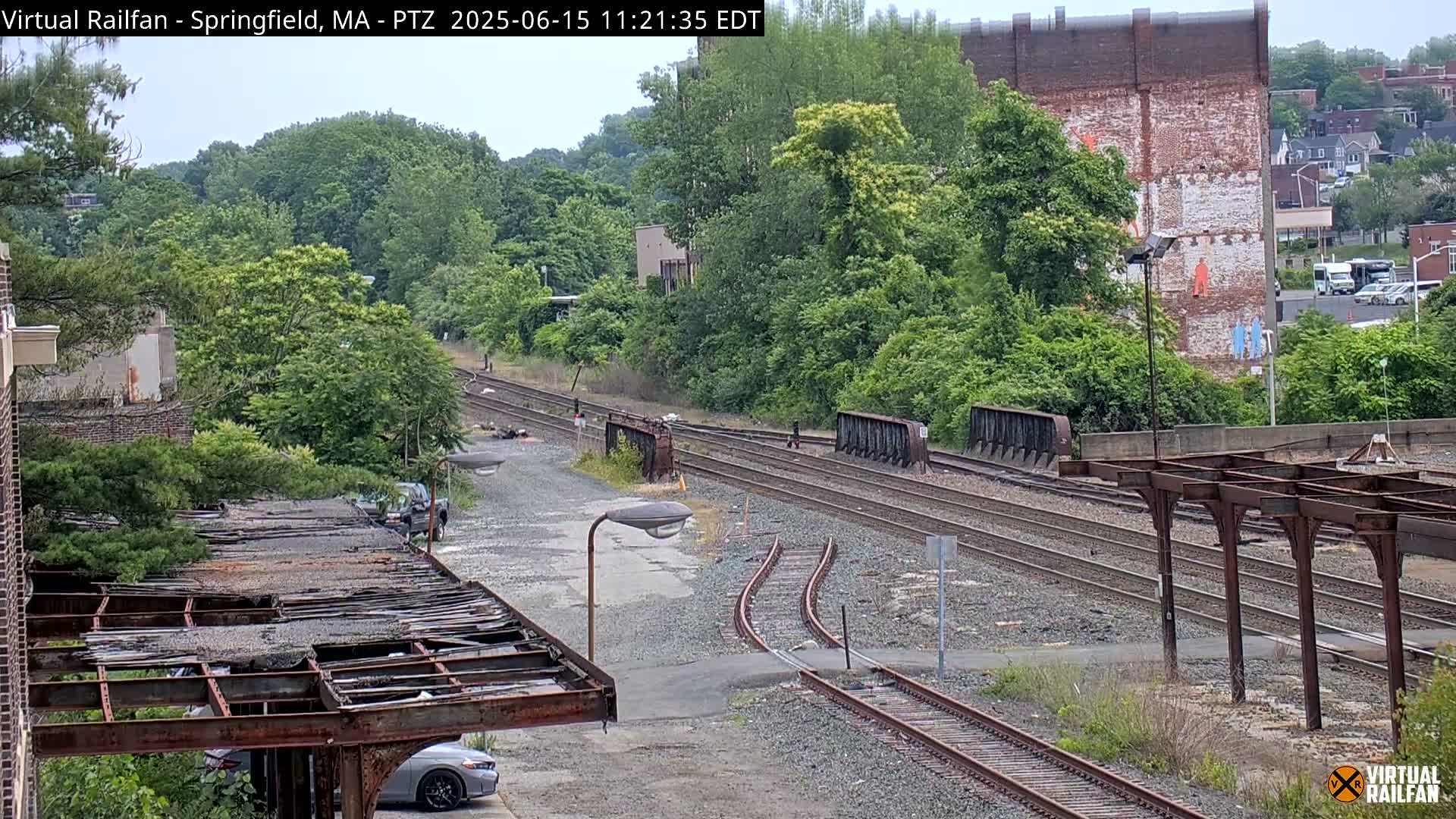 A view of railway tracks and a dilapidated structure under a mostly cloudy sky, with lush green trees and buildings in the background.
