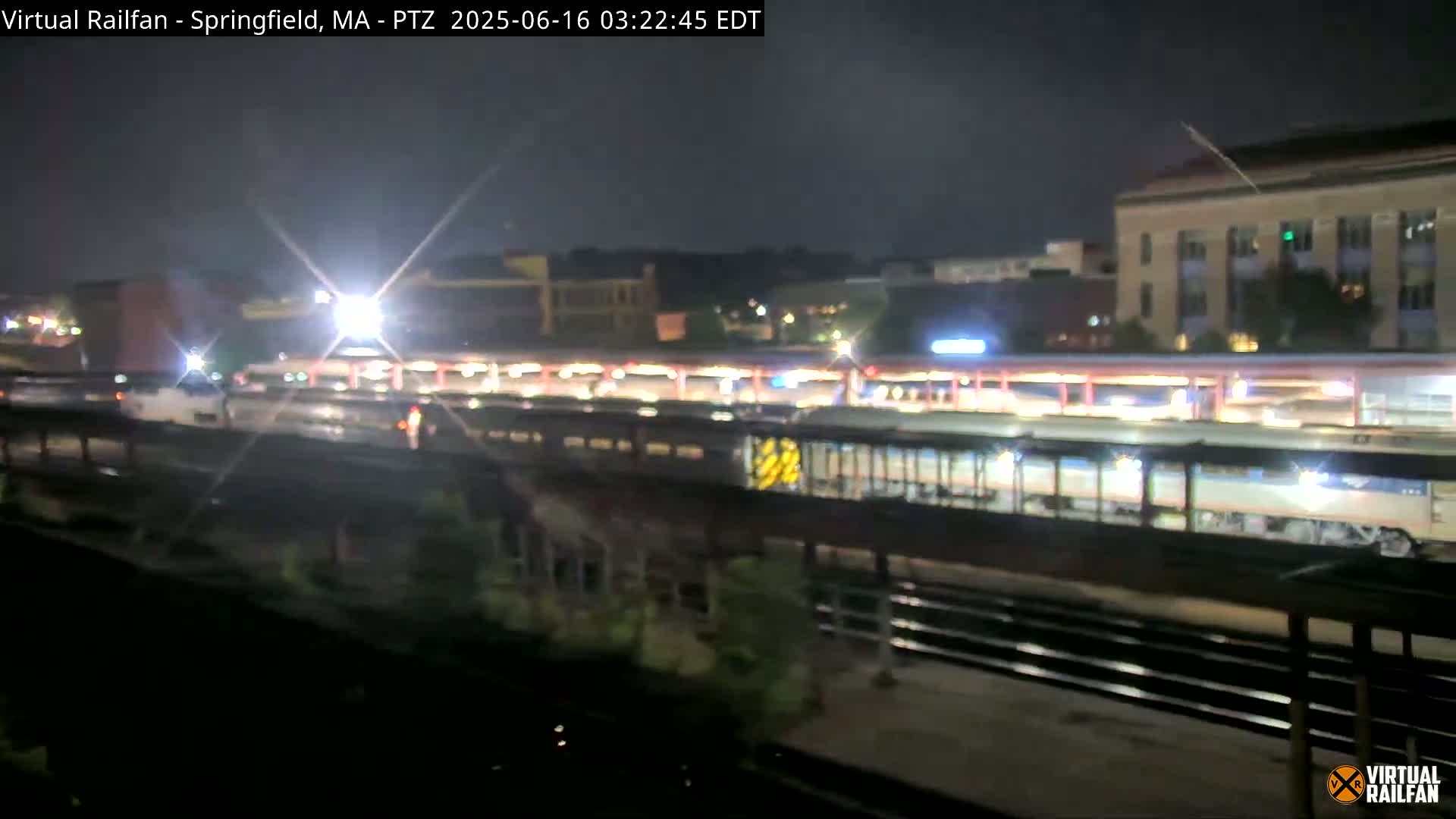 A nighttime view of a train at a station, with city buildings in the background under a dark sky.