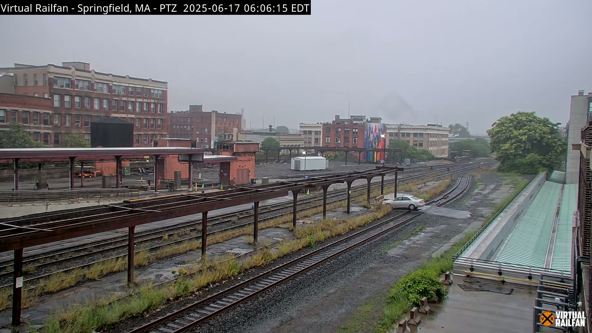 A rainy view of a train yard with several tracks, a platform, and industrial buildings in the background.