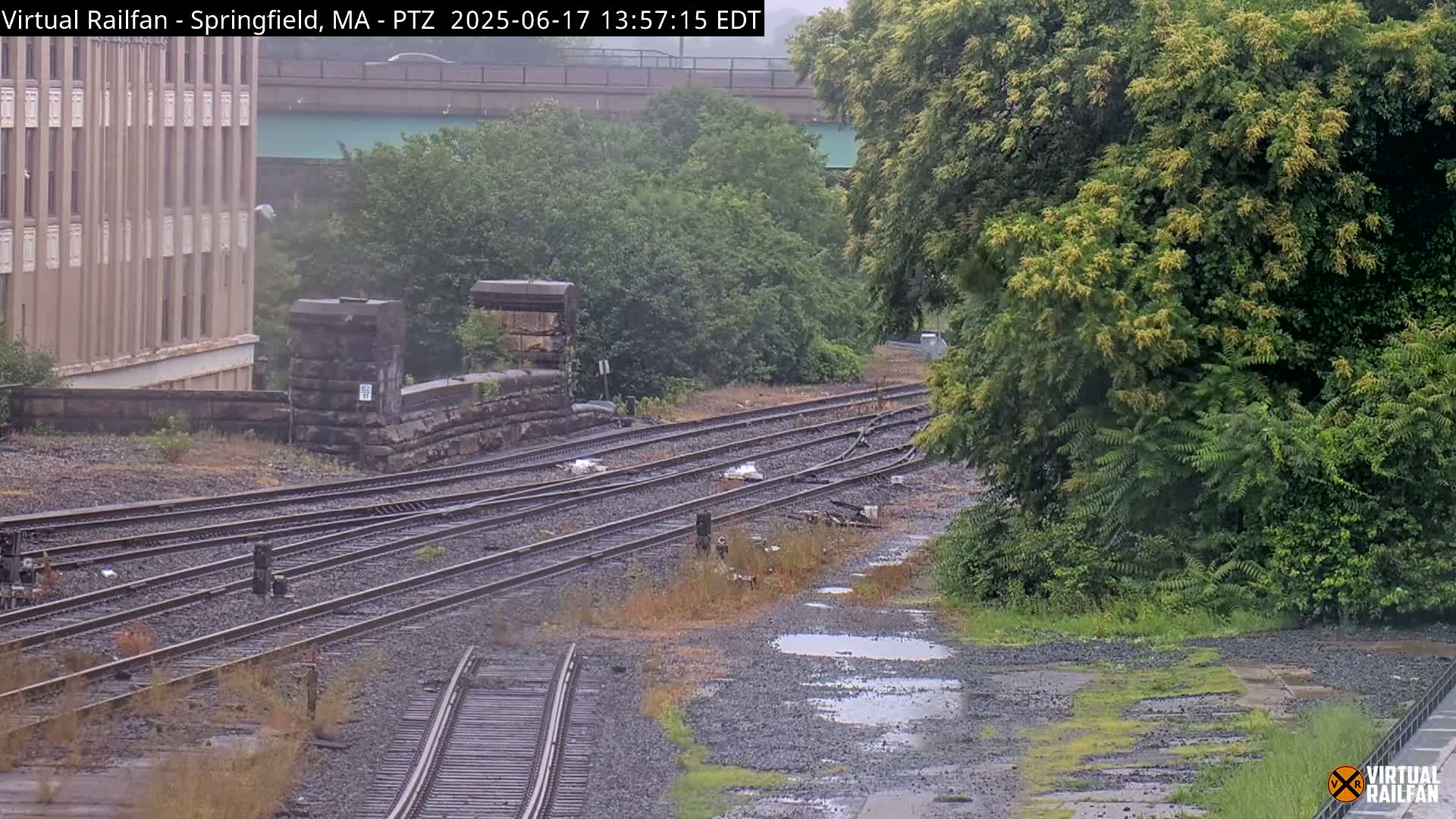 A view of several railway tracks on an overcast day with puddles on the ground next to a stone structure and overgrown vegetation.