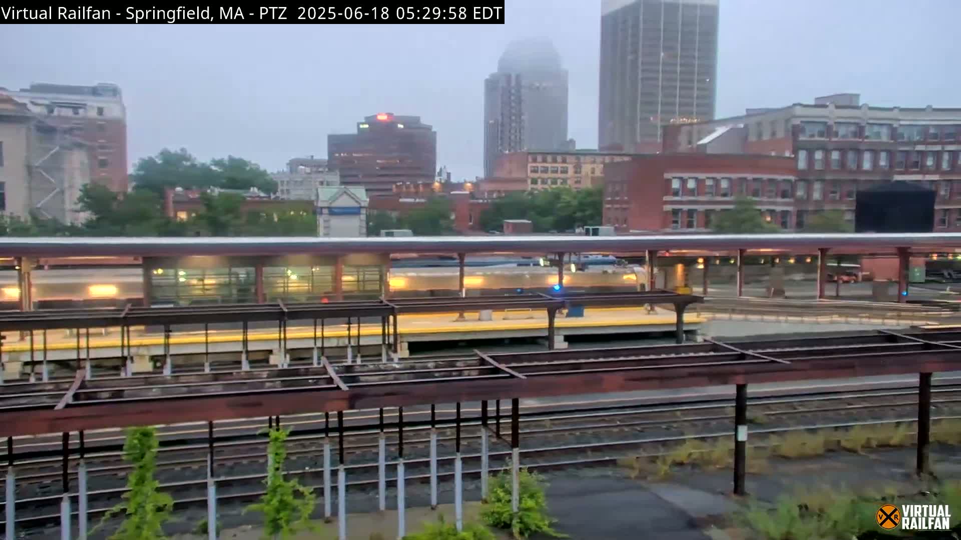 A train sits at a station platform in a city under an overcast sky.