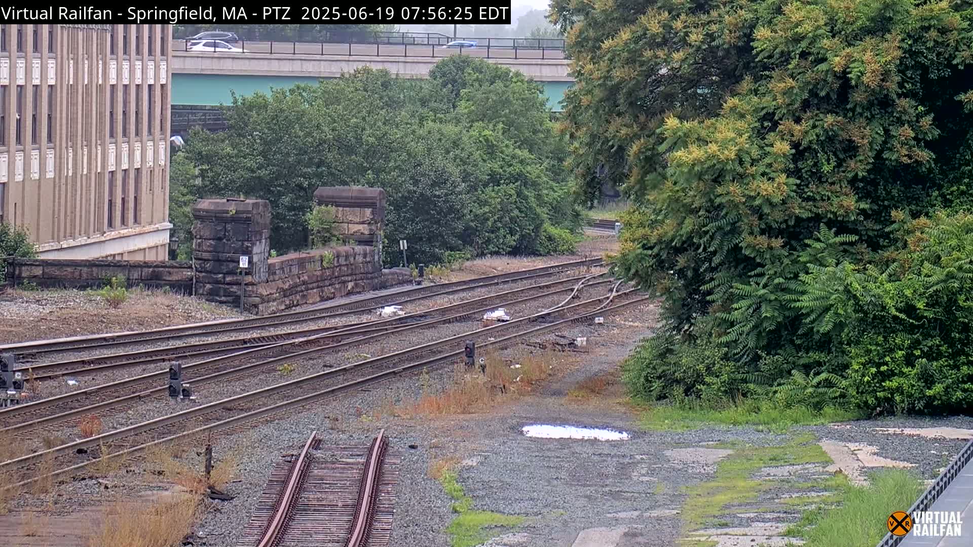 A high-angle view shows a railway switching area with several sets of tracks, a stone retaining wall, and lush green vegetation under an overcast sky.