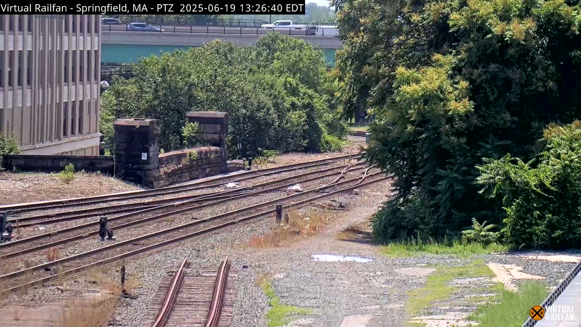 A high-angle view shows multiple train tracks that curve near a stone wall and lush green trees under a sunny sky.