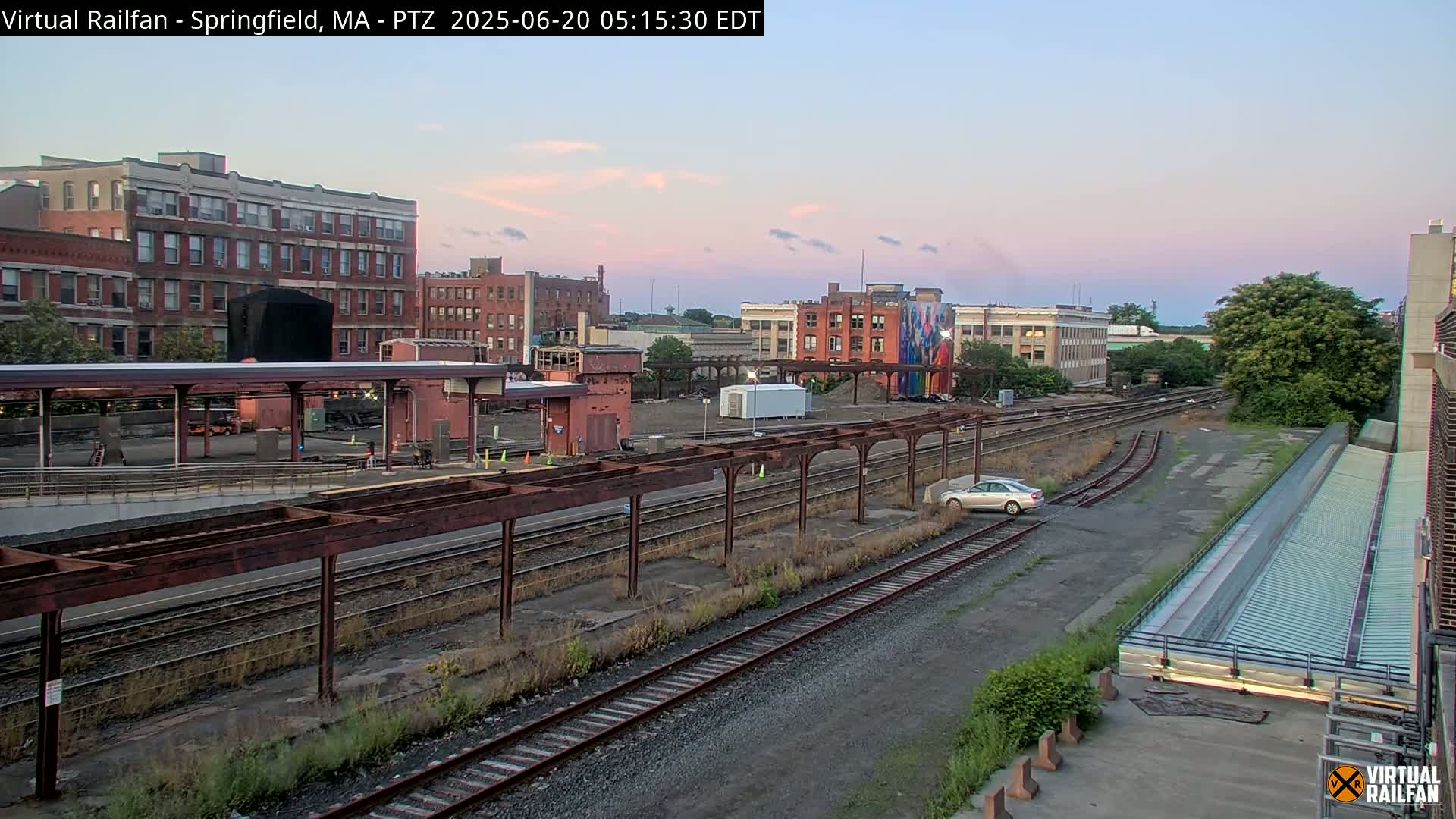 A train station with several railway tracks under a partly cloudy, dusky sky, surrounded by brick buildings and some vegetation.