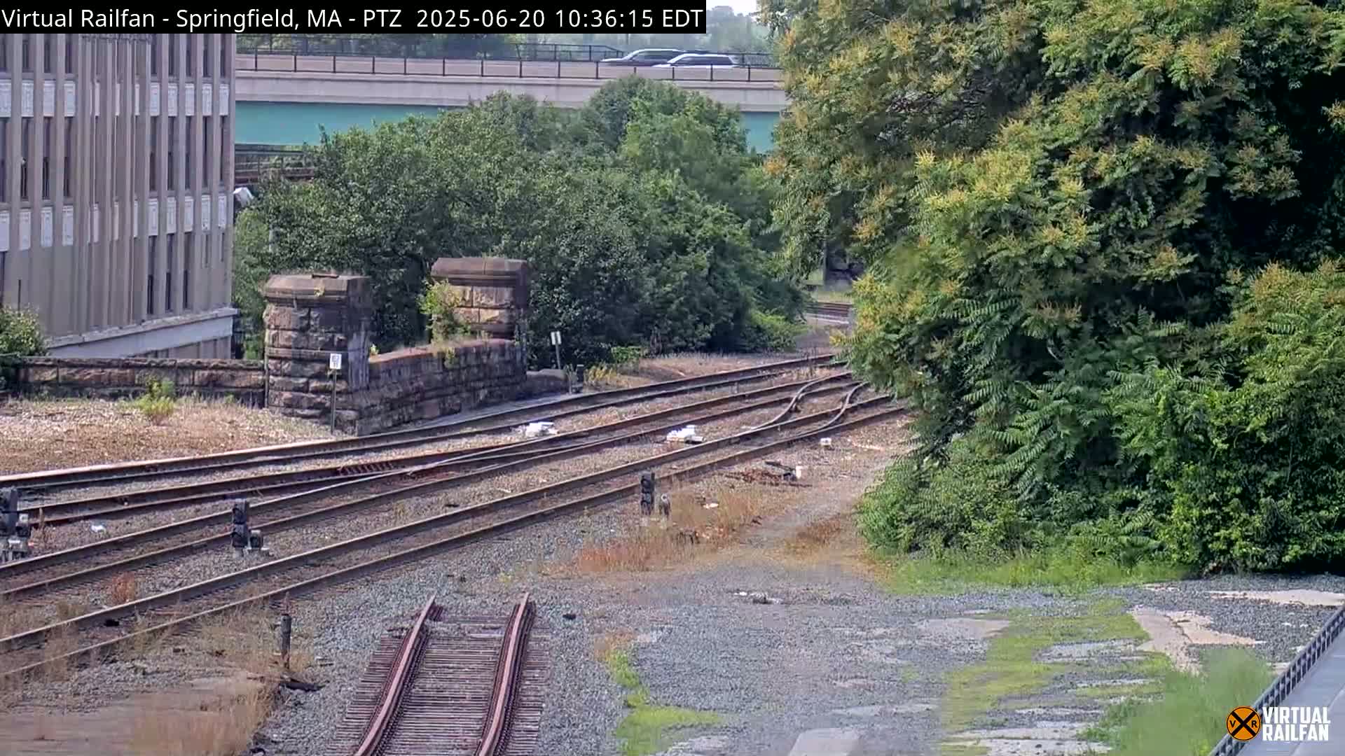 Several train tracks converge near a stone wall and lush green vegetation under a partly sunny sky.