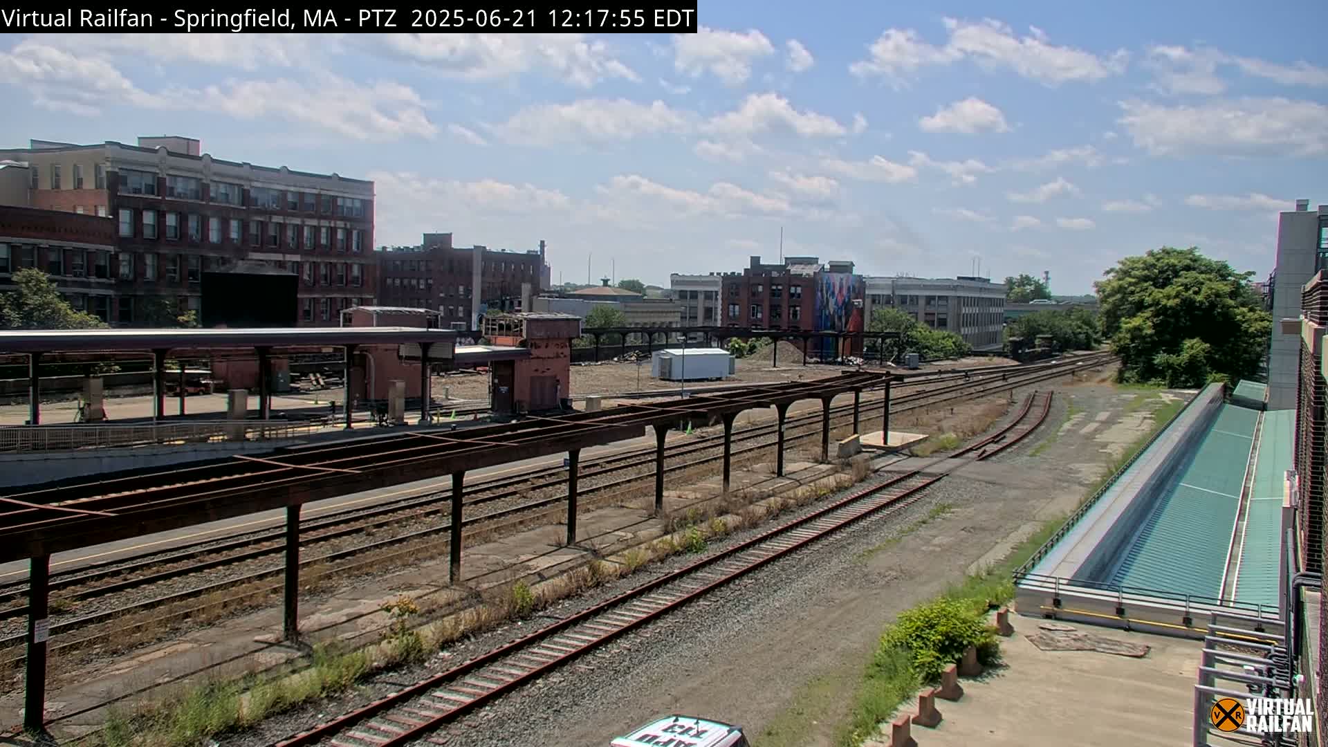 A partly sunny day reveals a train station with multiple tracks, platforms, and surrounding buildings under construction.