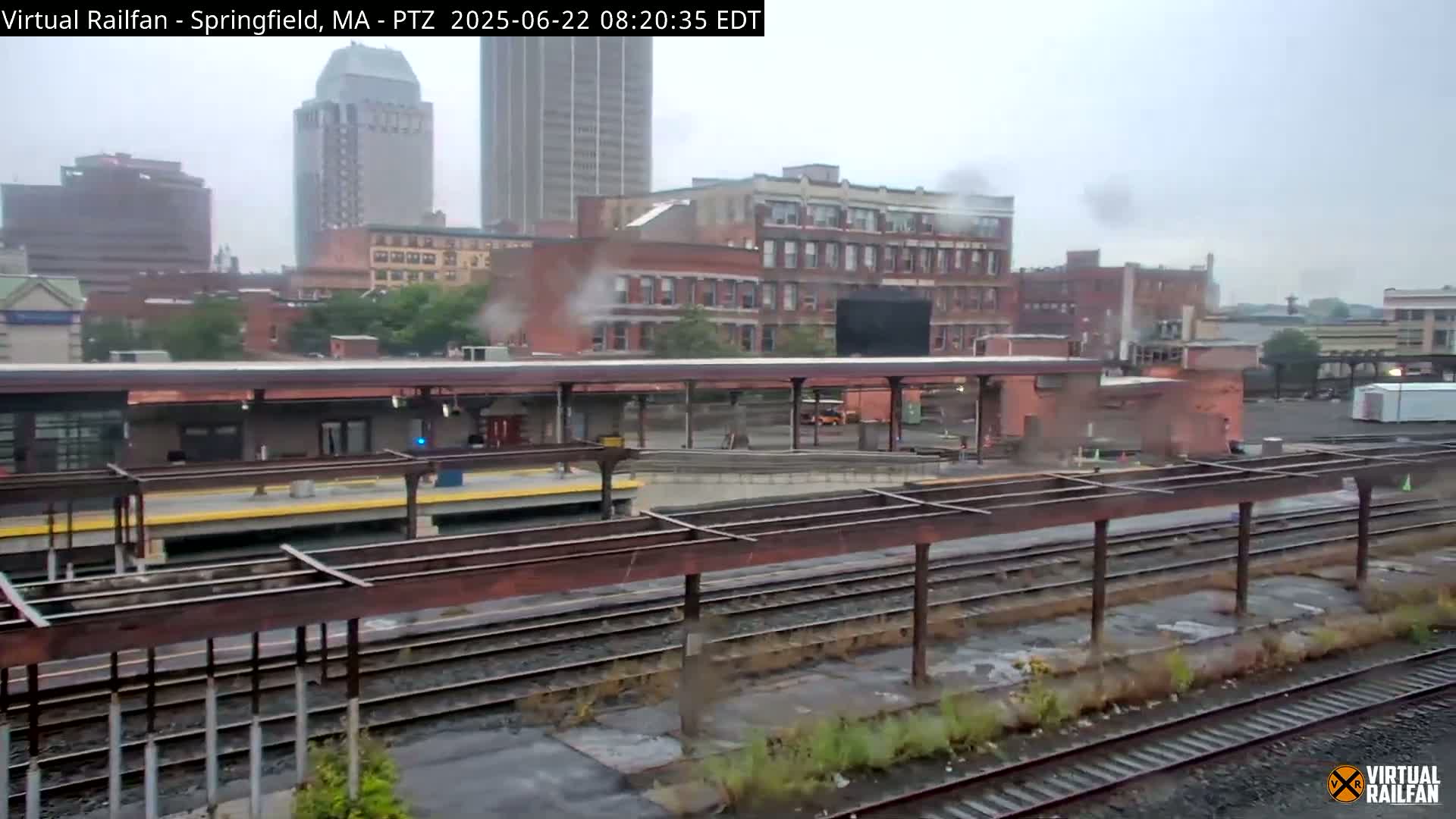 A train station platform with several tracks and buildings in the background under an overcast sky.