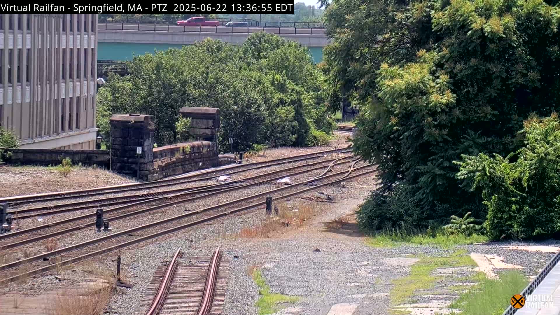 A high-angle view of a railway switching yard with multiple tracks, a stone retaining wall, and lush green trees under a sunny sky.