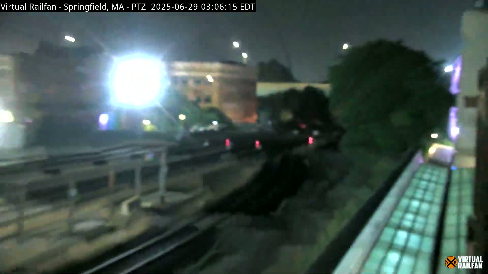A nighttime view from a slightly elevated position shows train tracks, parked cars, and buildings under mostly dark conditions with some lights visible.