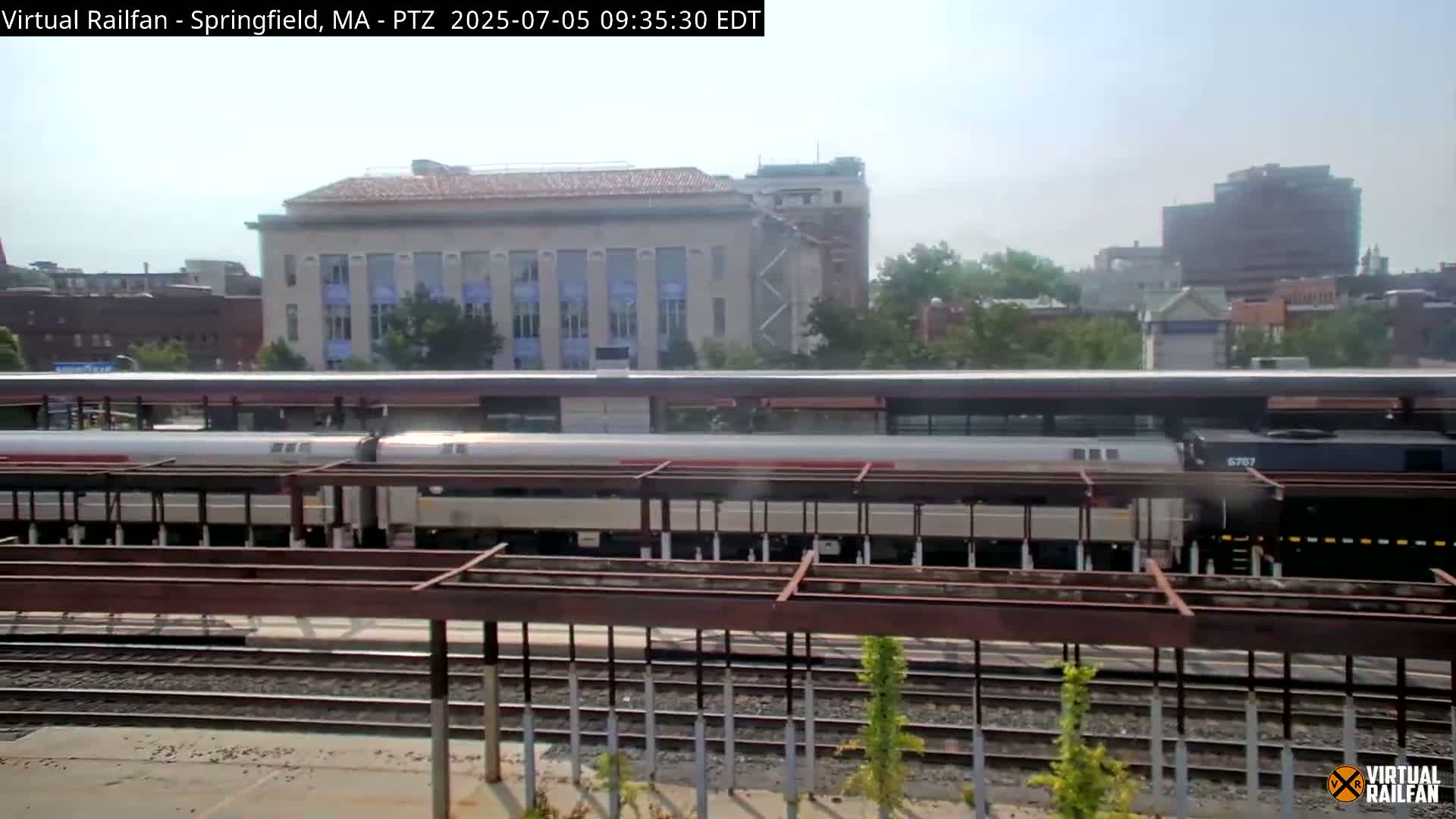 A passenger train is at a station on a partly sunny day, with buildings and trees visible in the background.