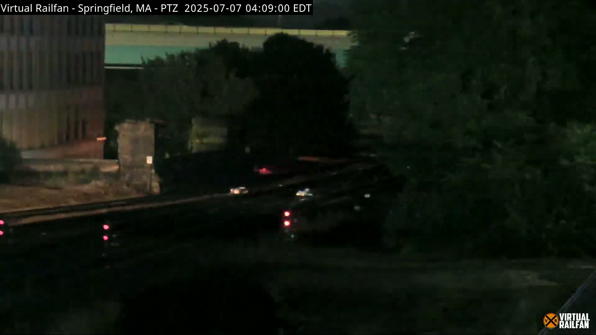 At night, a dark-colored vehicle is partially visible near a railway track, with a building and trees in the background.