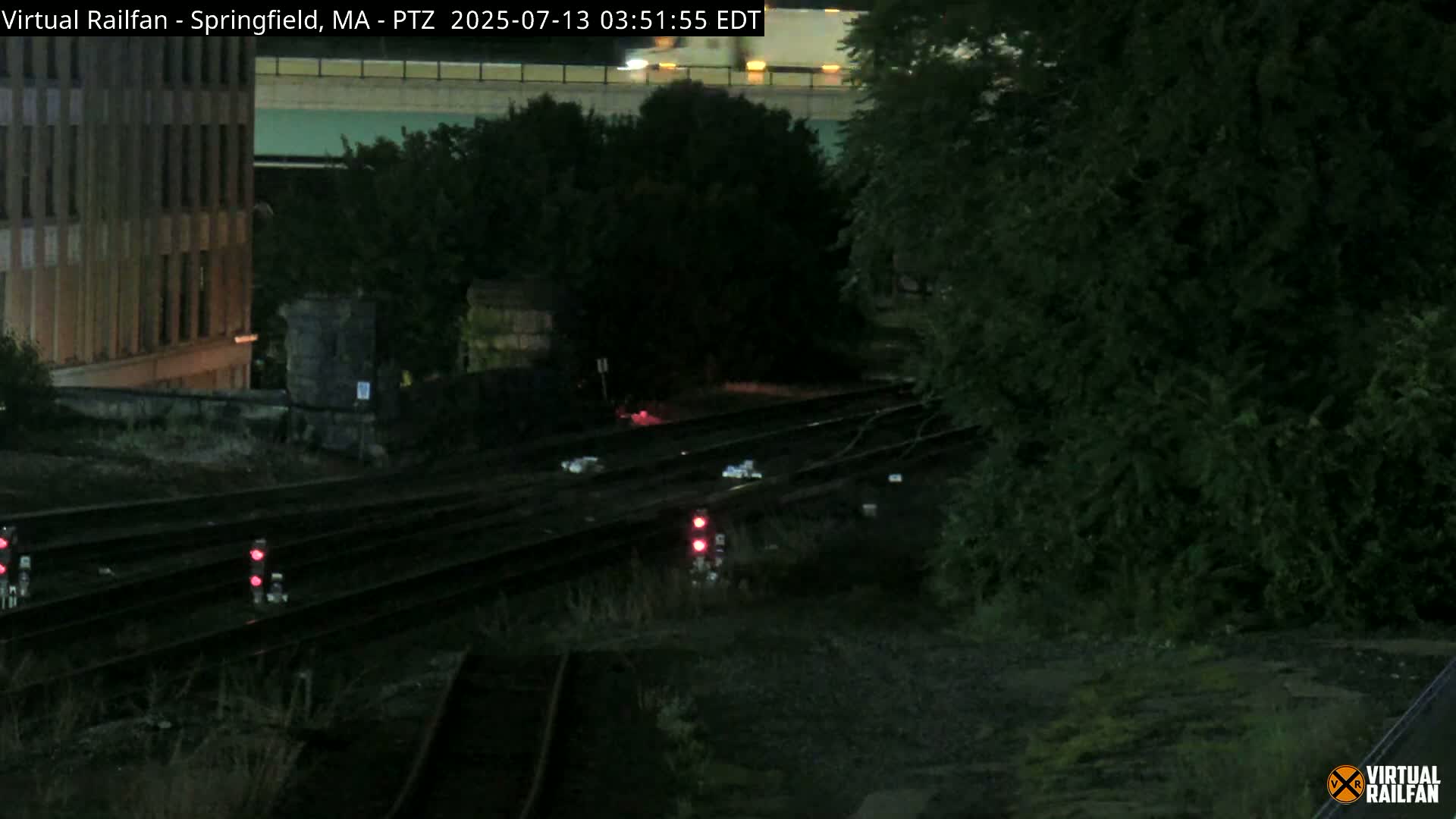 At night, a view of train tracks runs horizontally across the frame, near a building and lush, dark foliage.