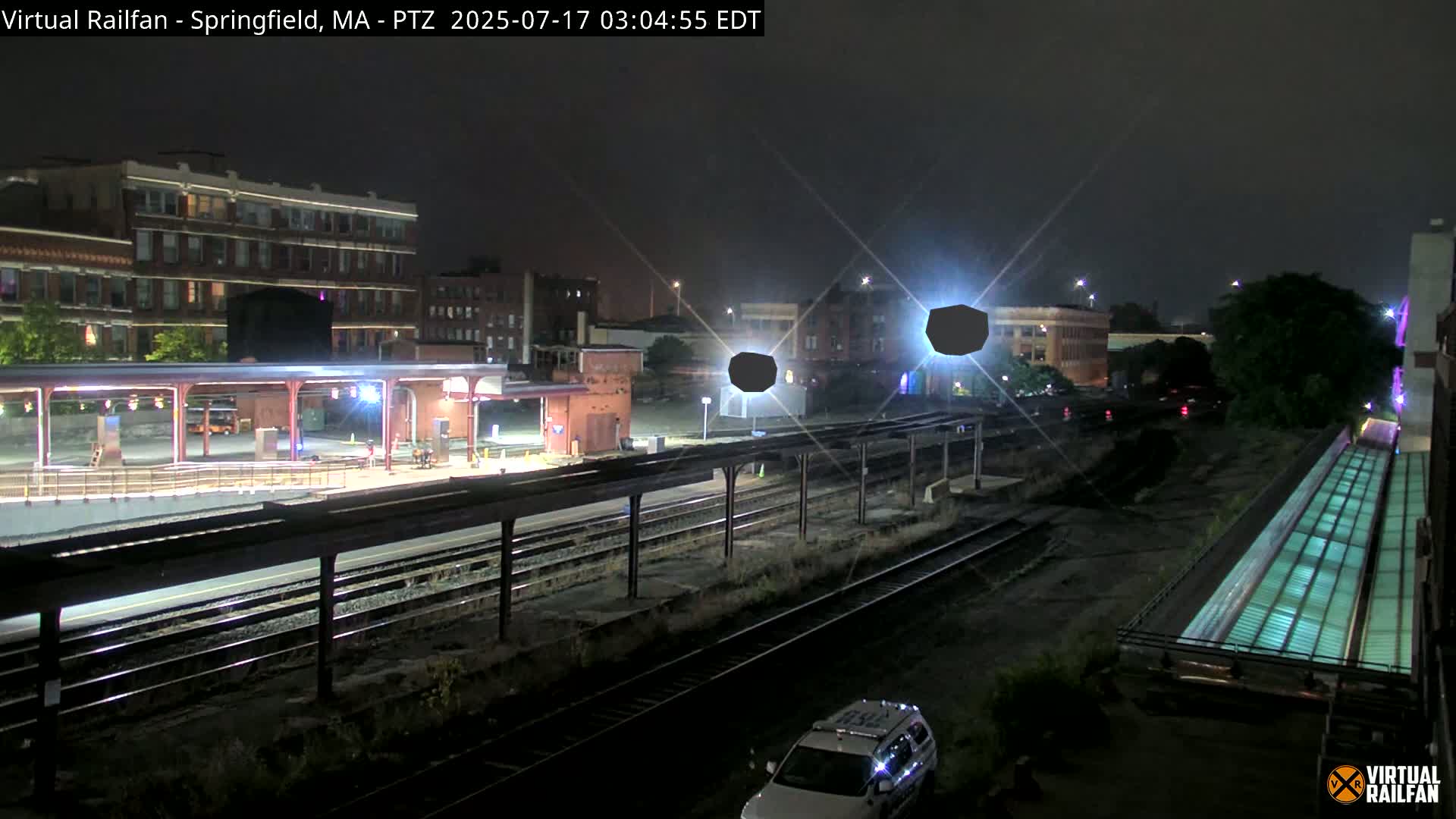 A nighttime view of a train station platform and tracks, with a police car visible in the foreground, under partly cloudy skies.