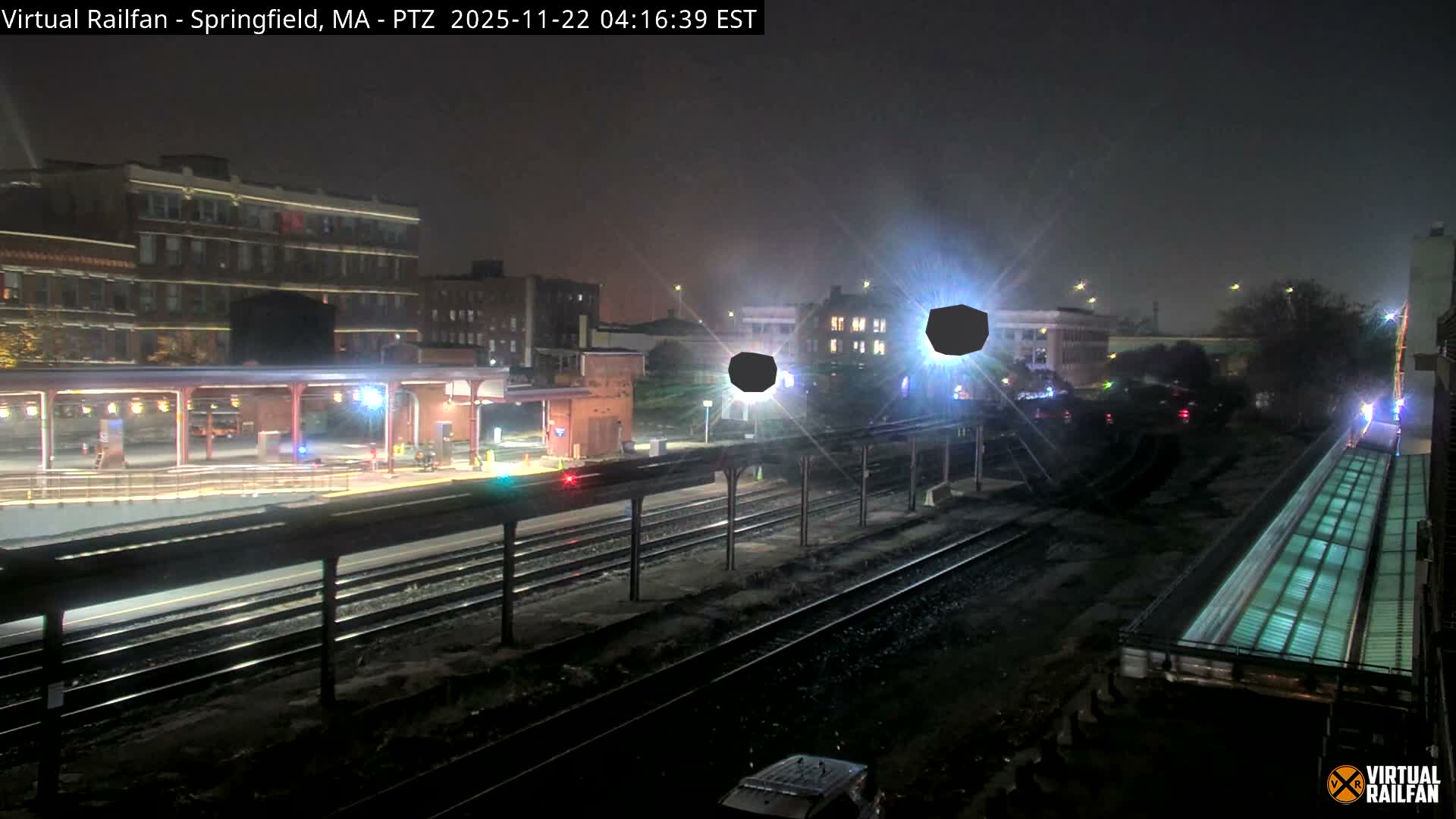 An overhead nighttime view depicts a train station with multiple tracks and brightly lit covered platforms, surrounded by urban buildings under an overcast sky.