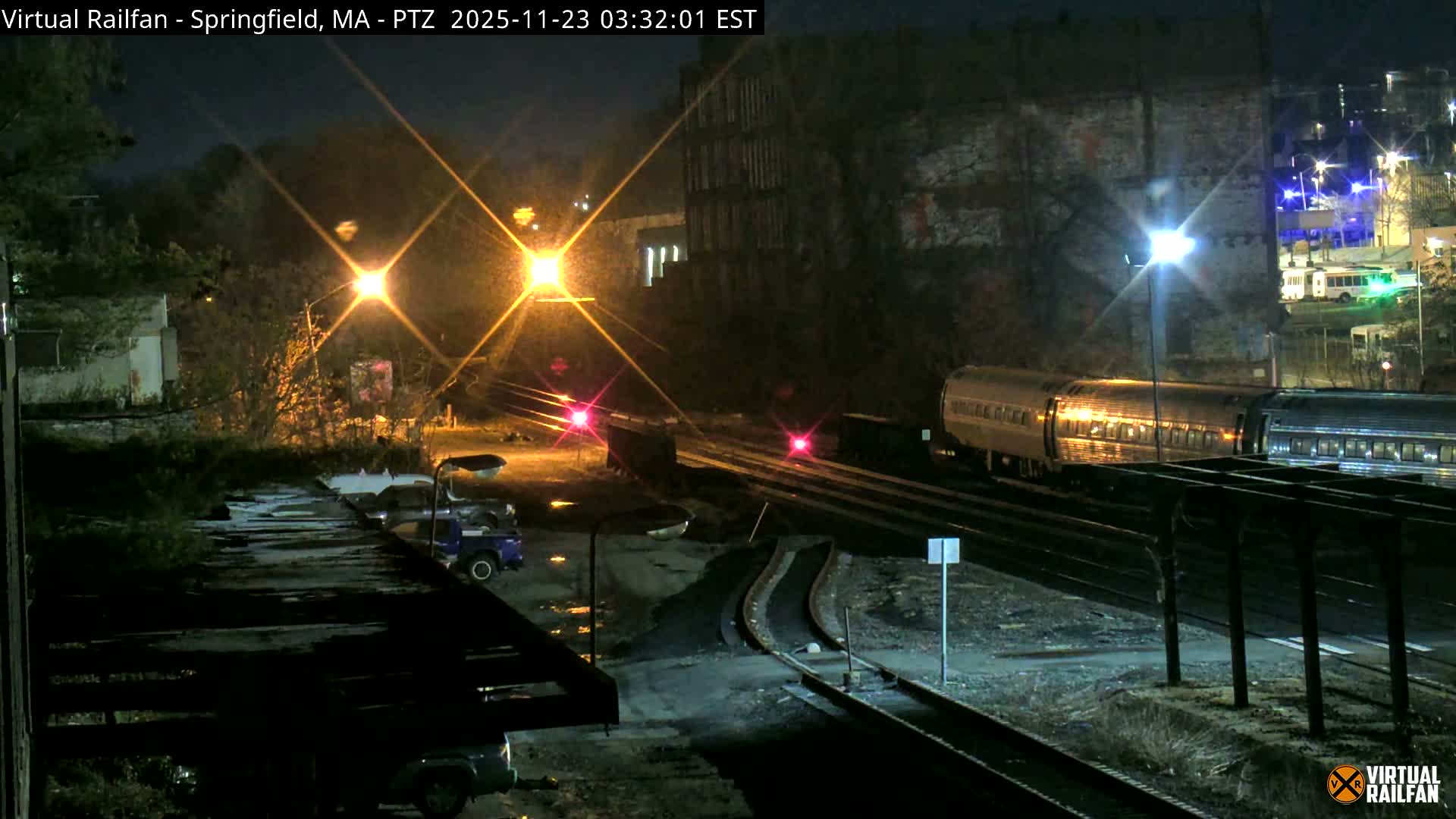A passenger train with reflective cars moves along multiple tracks at night, illuminated by bright streetlights that cast lens flares and reflect off the wet ground, indicating recent rain.