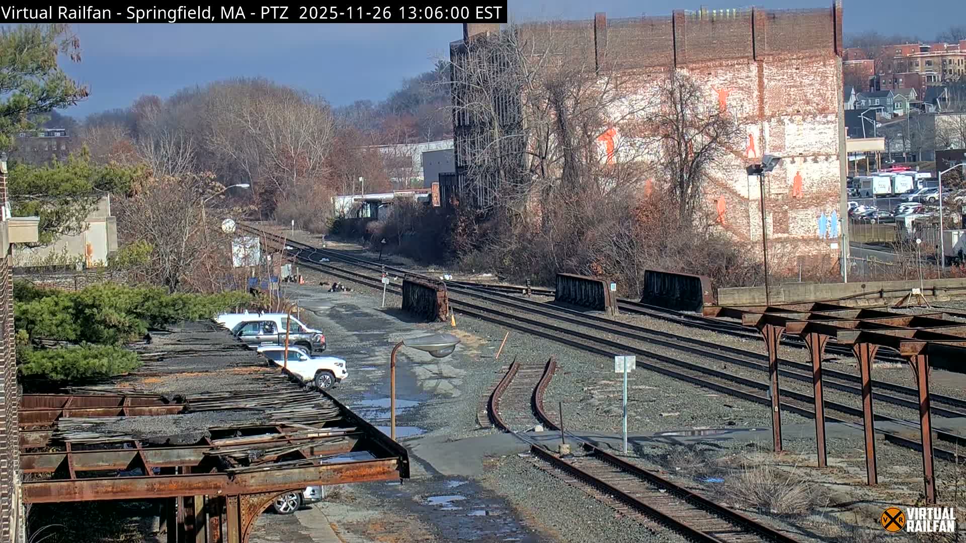 On a clear and sunny day, multiple railway tracks cut through an industrial landscape featuring a dilapidated structure with parked cars in the foreground, an old brick building with painted figures in the midground, and bare trees on hills in the background.