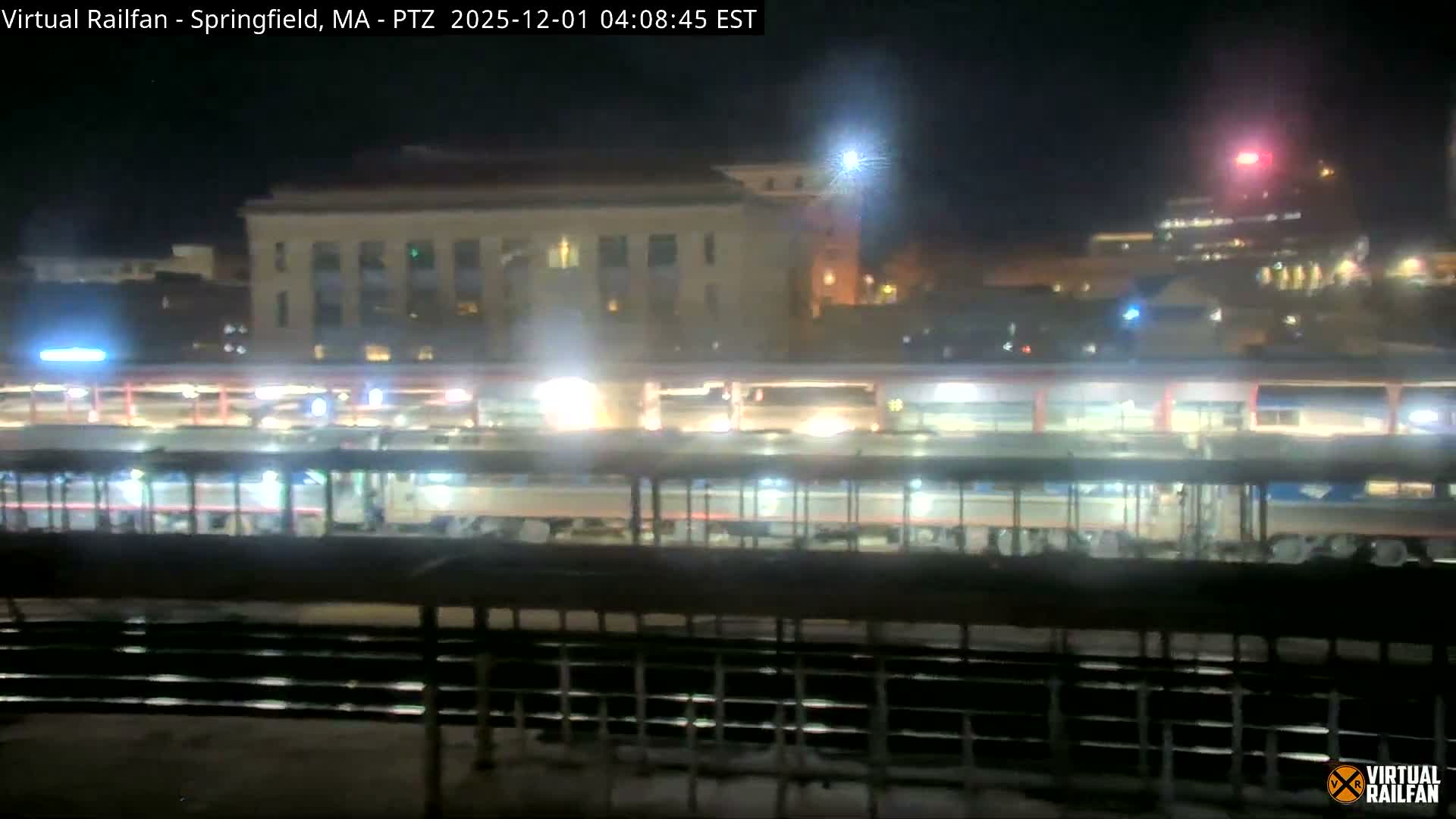 A hazy nighttime scene reveals brightly lit train station platforms above multiple train tracks, with illuminated city buildings visible in the background.
