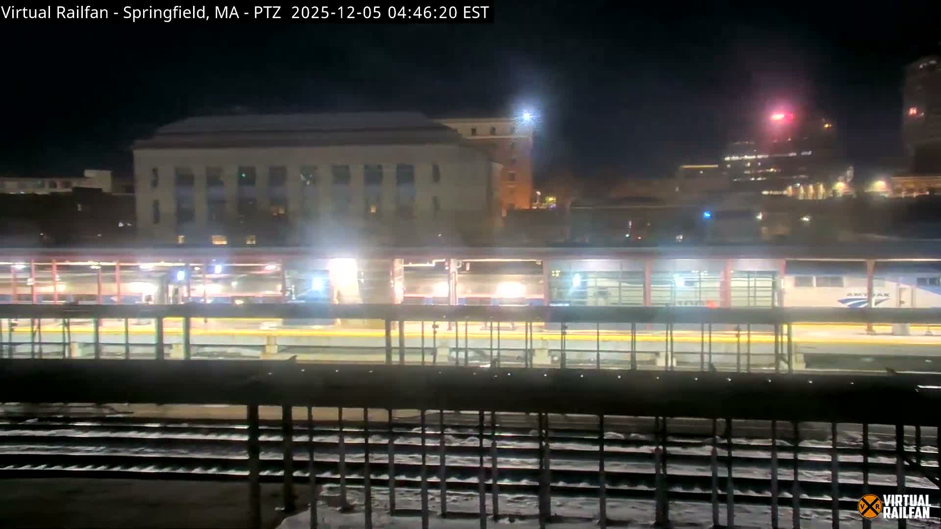 A nighttime view captures an Amtrak train parked at a brightly lit platform above snow-dusted tracks, with city buildings visible in the background under a clear, dark sky.