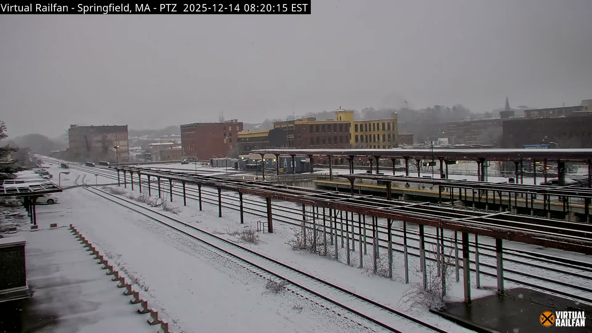 A nighttime view captures an Amtrak train parked at a brightly lit platform above snow-dusted tracks, with city buildings visible in the background under a clear, dark sky.