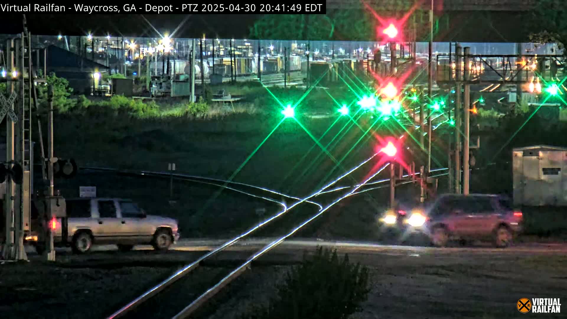 At night, a silver SUV and a dark-colored SUV drive past a railroad crossing with flashing red and green lights.