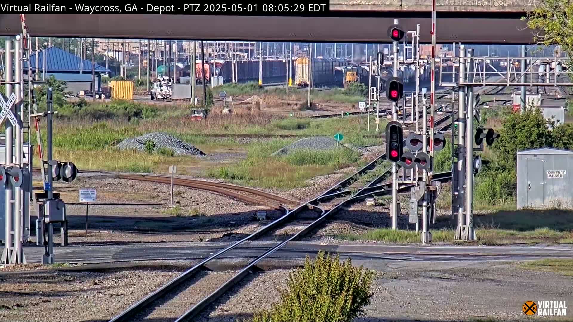 A sunny day at a railroad yard shows a complex intersection of train tracks with several signals and a rail crossing, alongside some industrial buildings and vegetation.