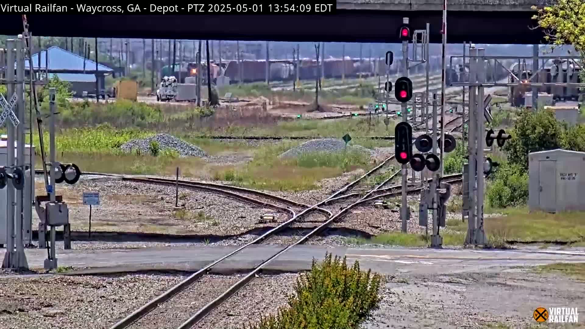 A sunny outdoor scene shows a railway intersection with multiple tracks converging, train signals displaying red lights, and some vegetation and small buildings in the background.