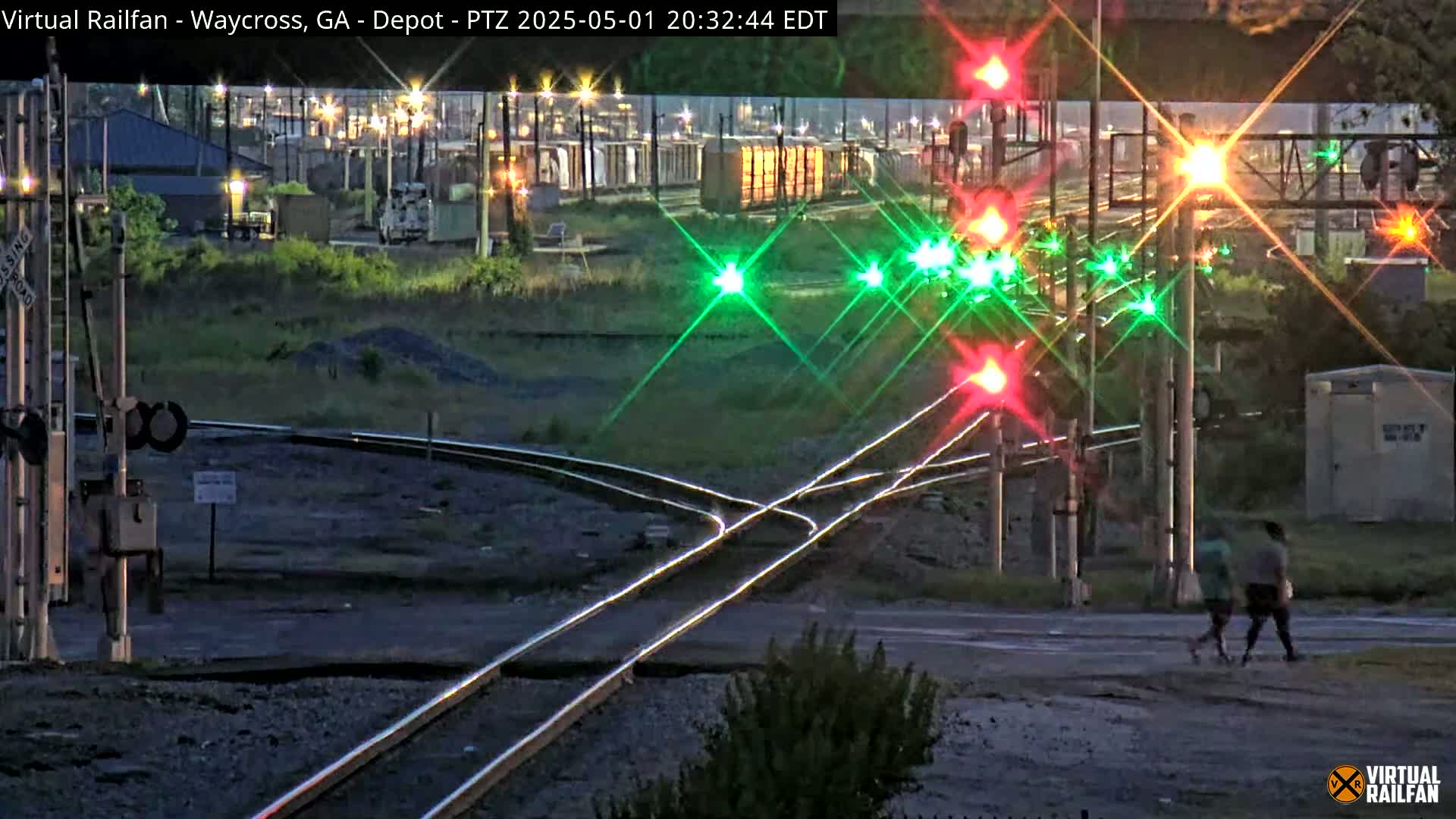 Two people walk across a road at a nighttime railroad crossing with active lights and signals, near an industrial area.
