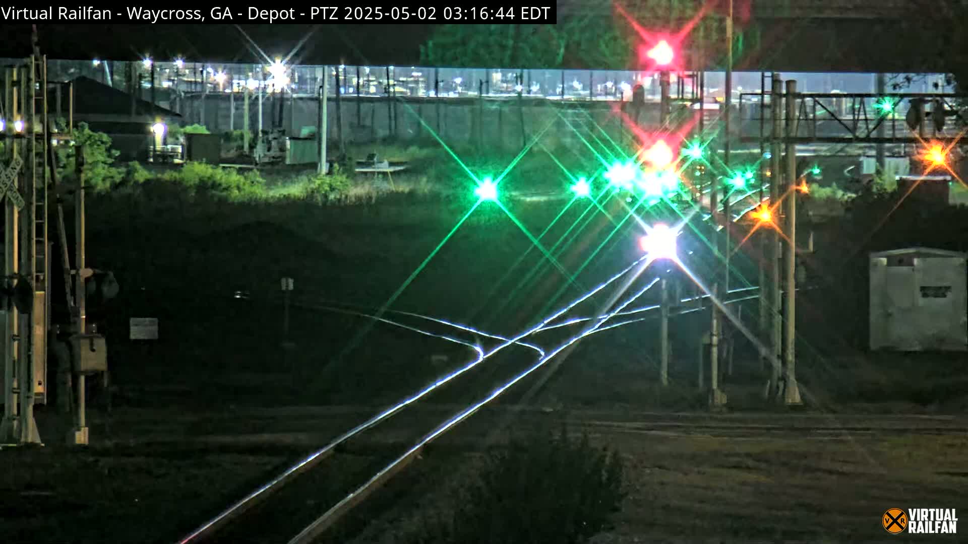 At night, a nighttime view shows railroad tracks and signals with some lights on, all under a dark sky.