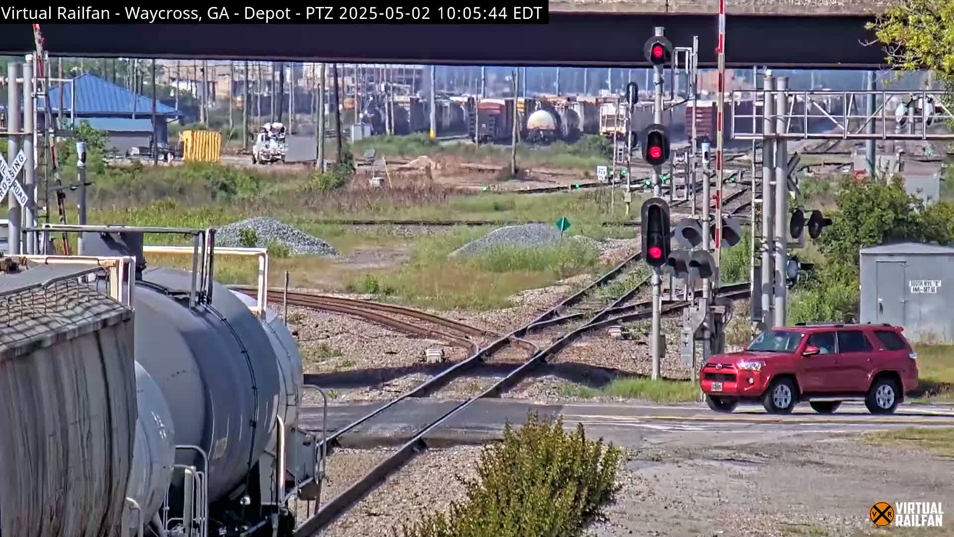A red SUV drives across a railroad crossing as a train of tank cars sits on the tracks under a sunny sky.