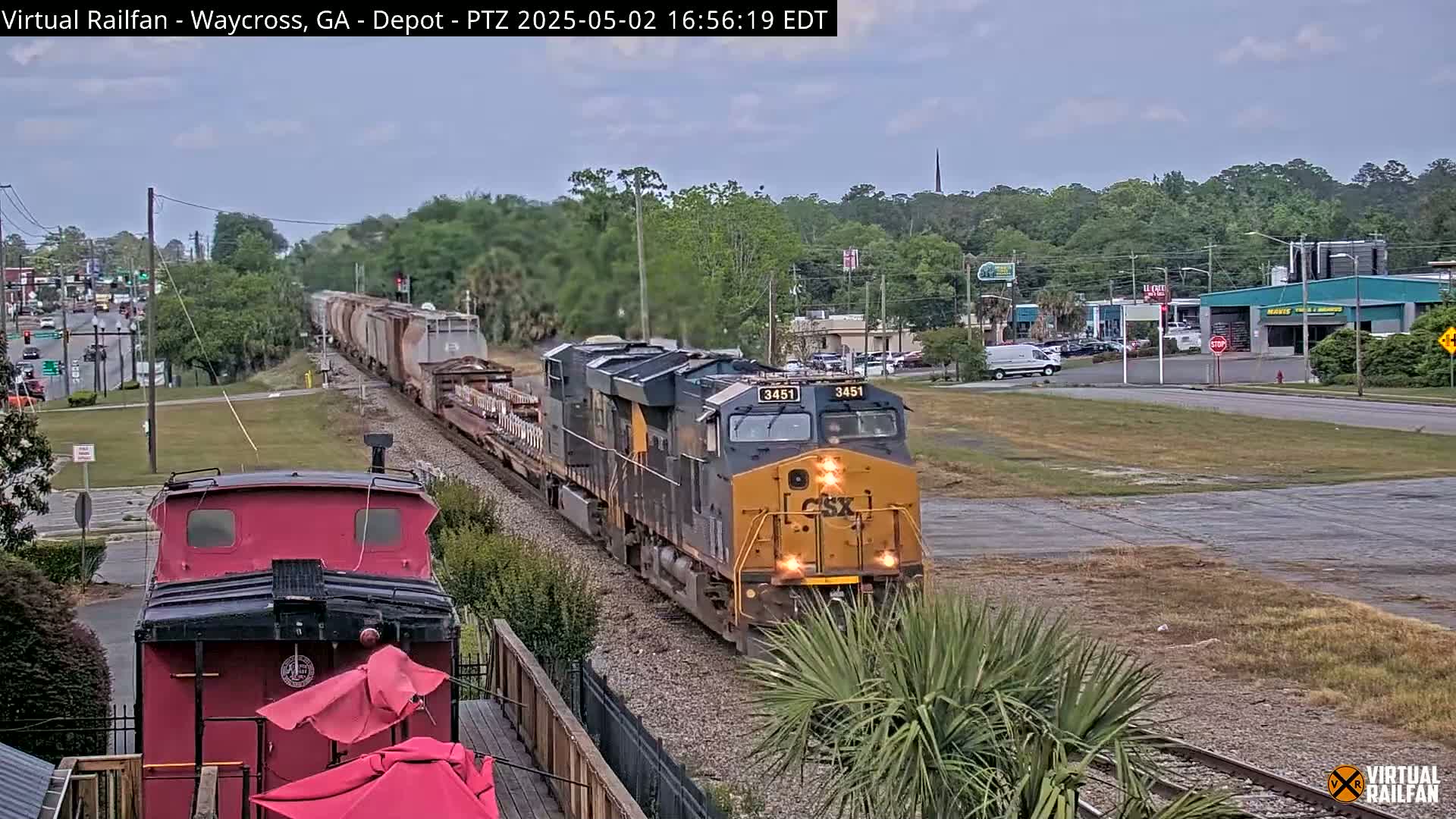 A long freight train passes through a town under an overcast sky, with a portion of a red caboose visible in the foreground.