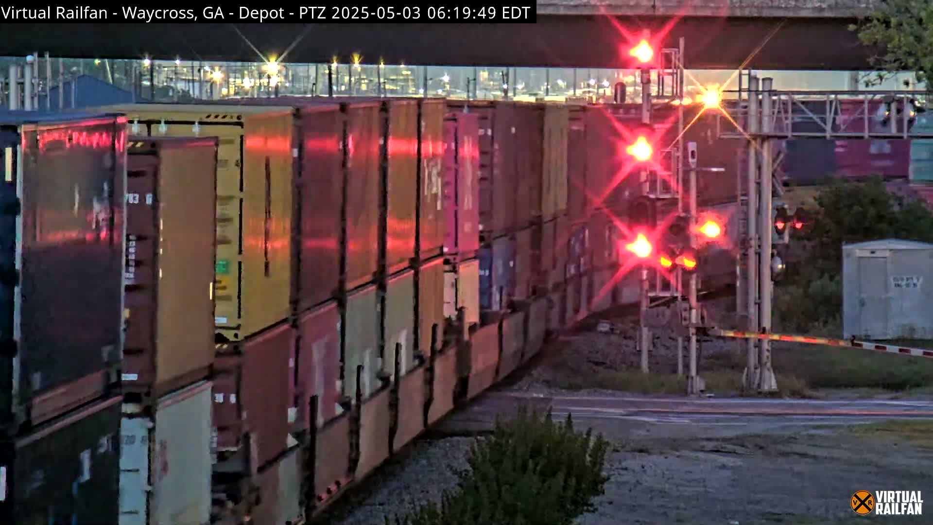 A long freight train passes under a railway crossing with flashing red lights at dusk.