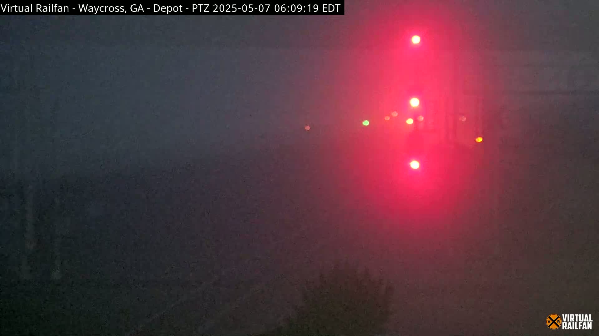 A foggy scene at night shows a set of red railroad signal lights illuminating the hazy atmosphere.