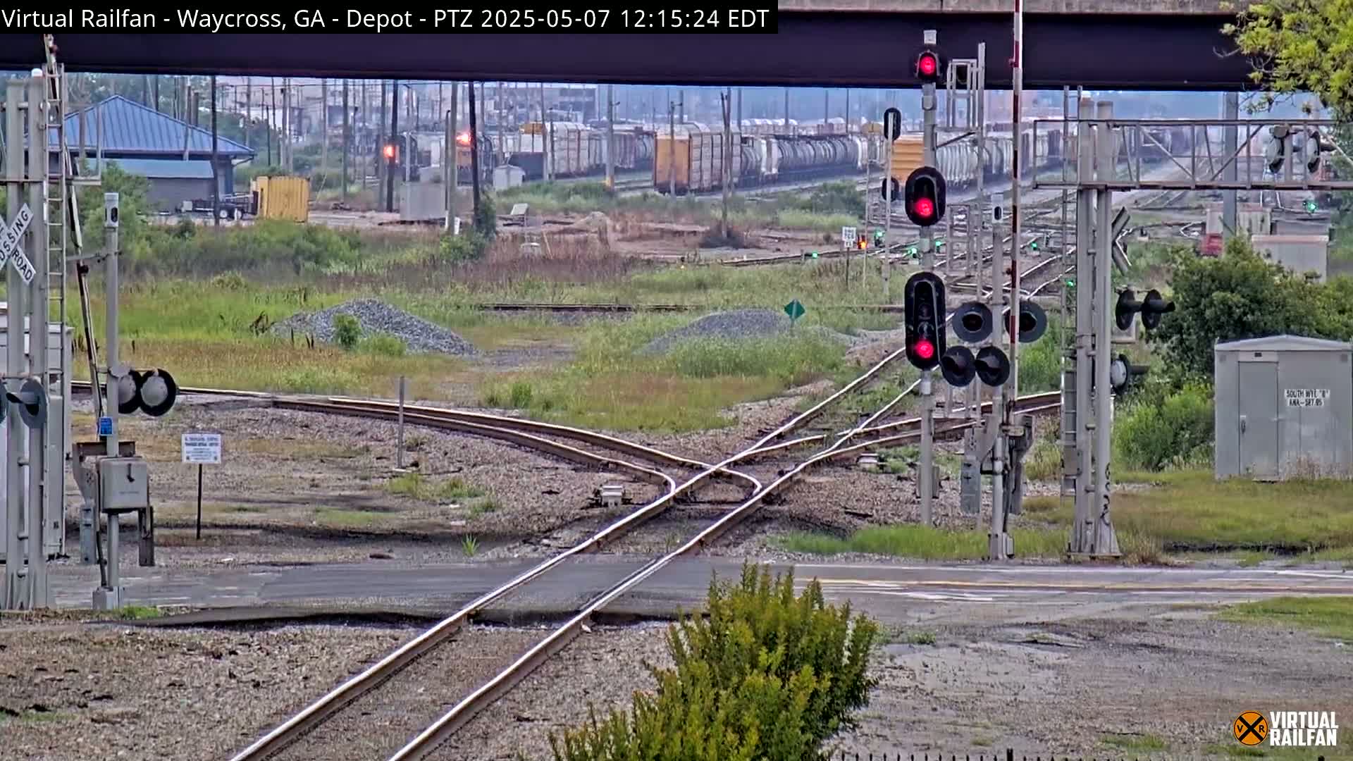 A train crossing with several sets of railroad tracks, signals showing red, and a freight train in the distance under an overcast sky.